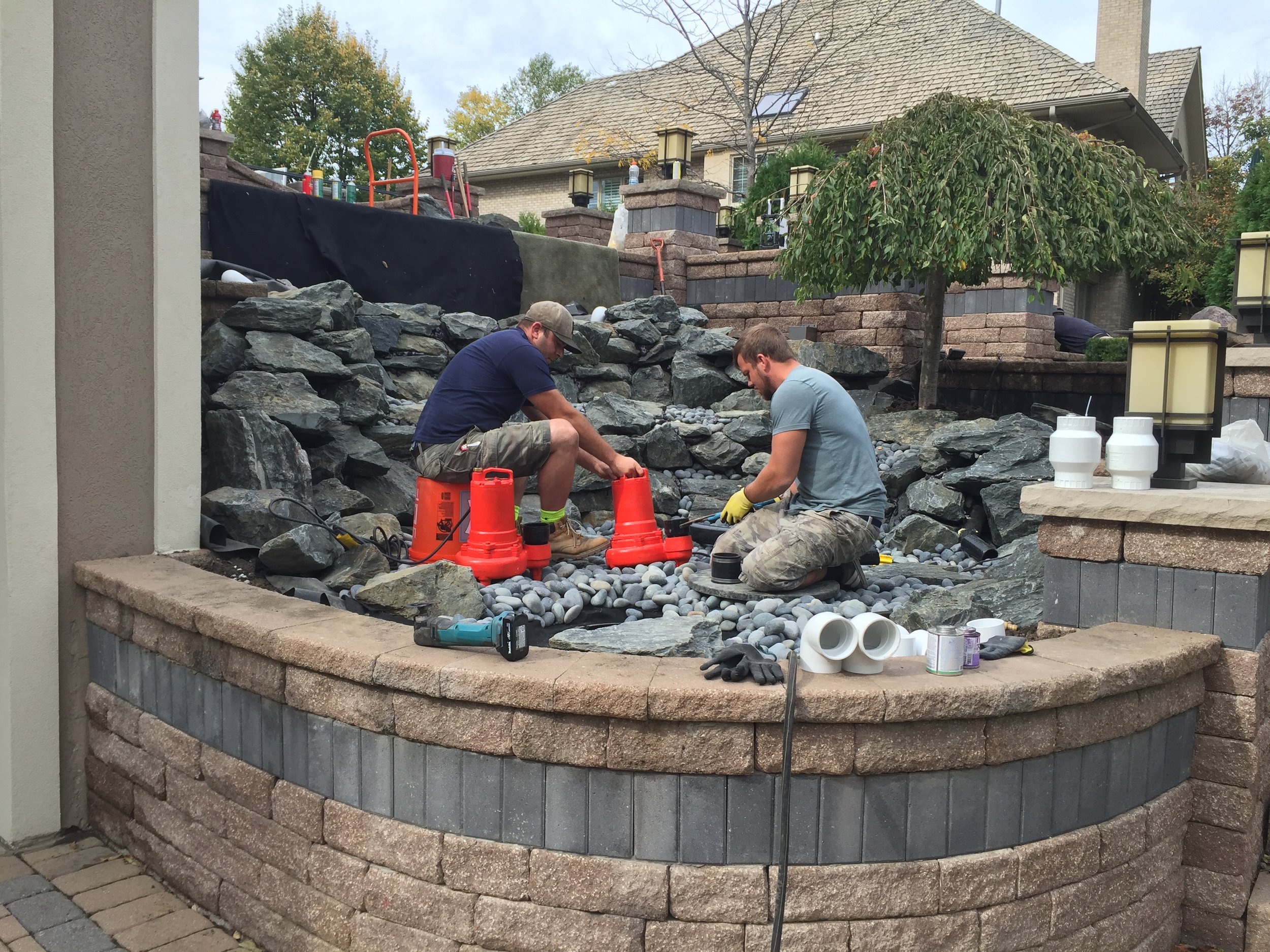 Two men working on a rock installation in a landscaped backyard. They are sitting on and around orange buckets among stacked stones and gravel. A house and trees are visible in the background.
