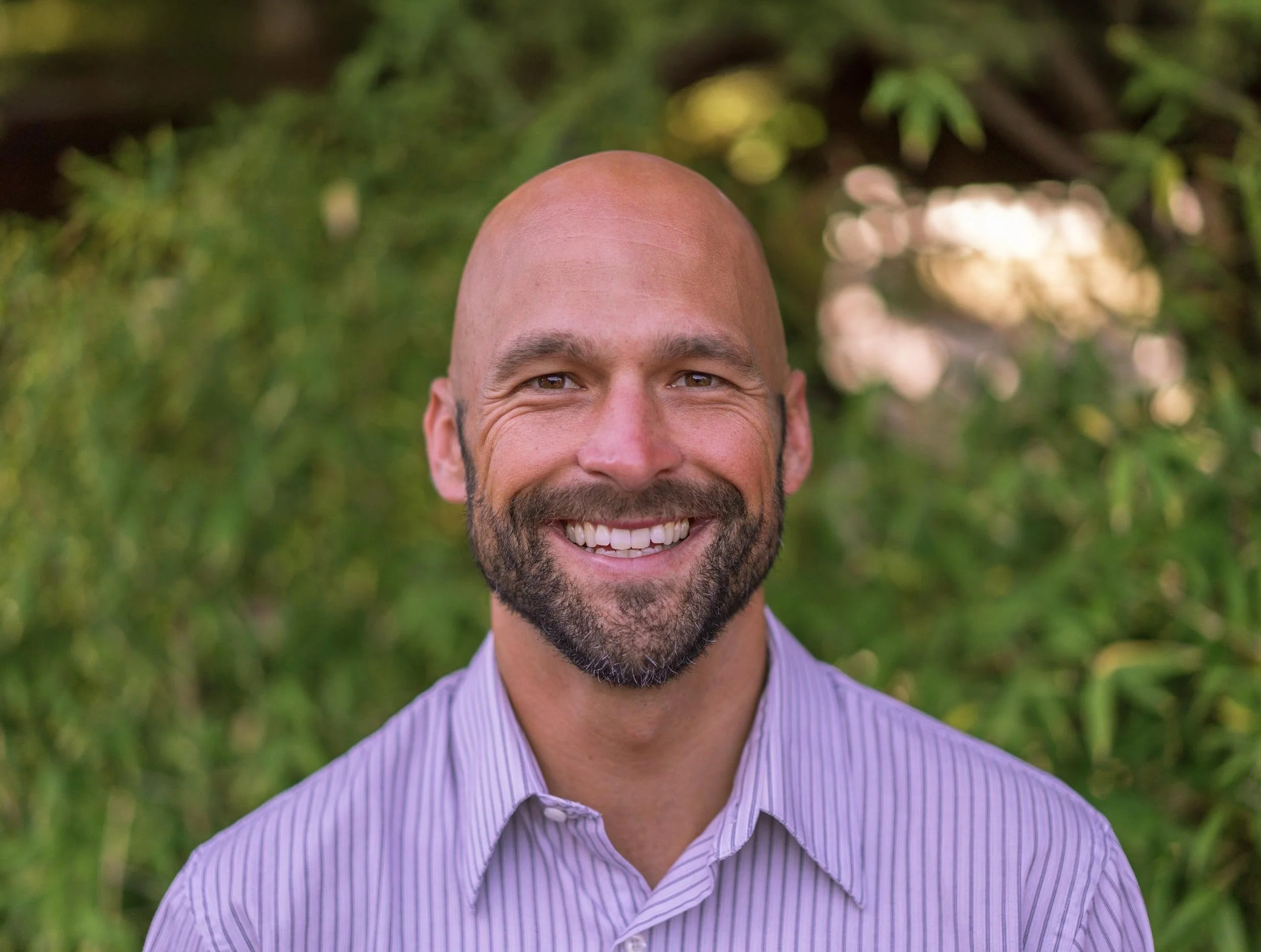 Dr. Daniel Madden with a beard, wearing a striped shirt, outdoor background with greenery.