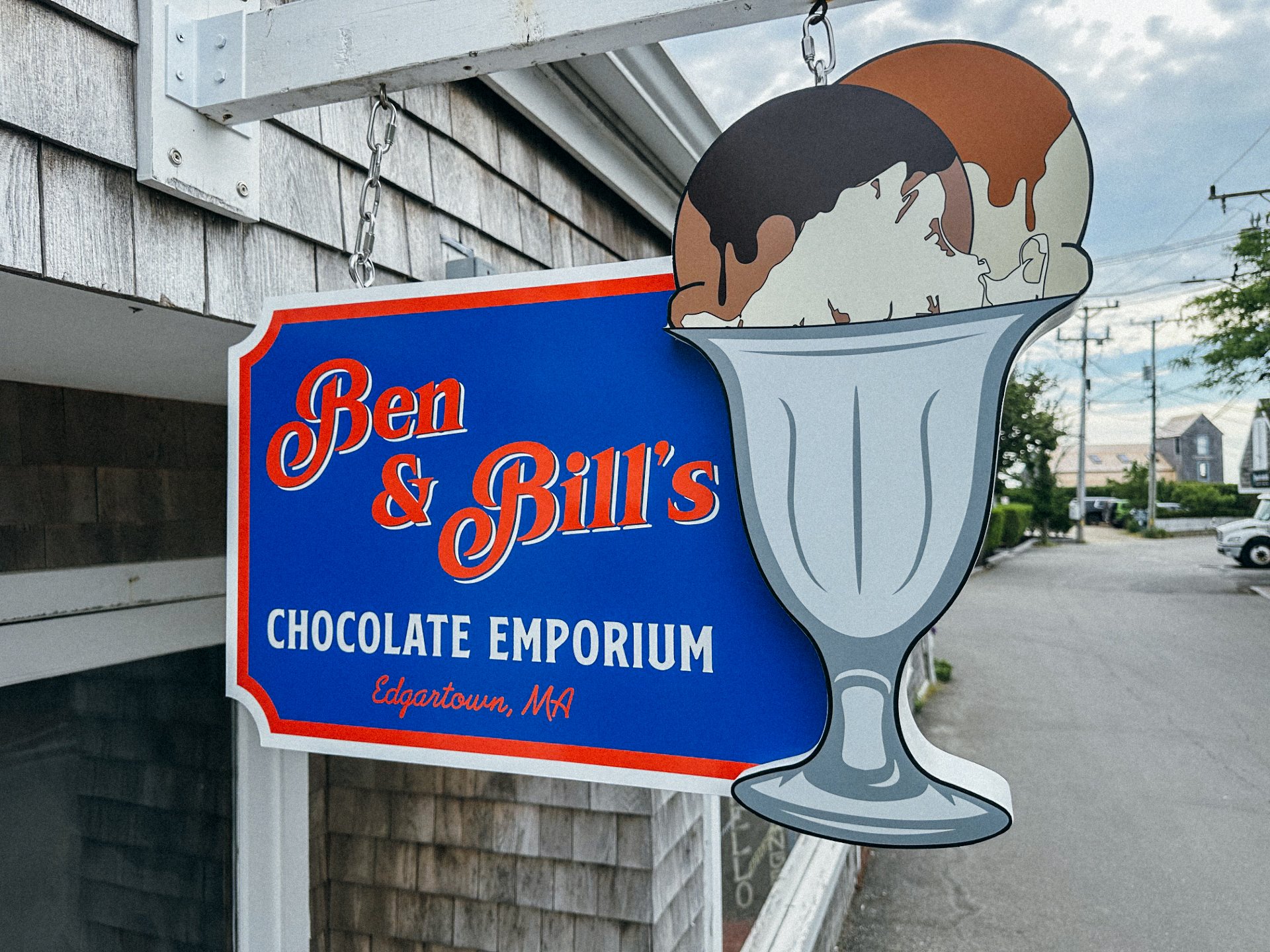 Sign for Ben & Bill's Chocolate Emporium with a large ice cream cone illustration hanging from a chain in front of a building