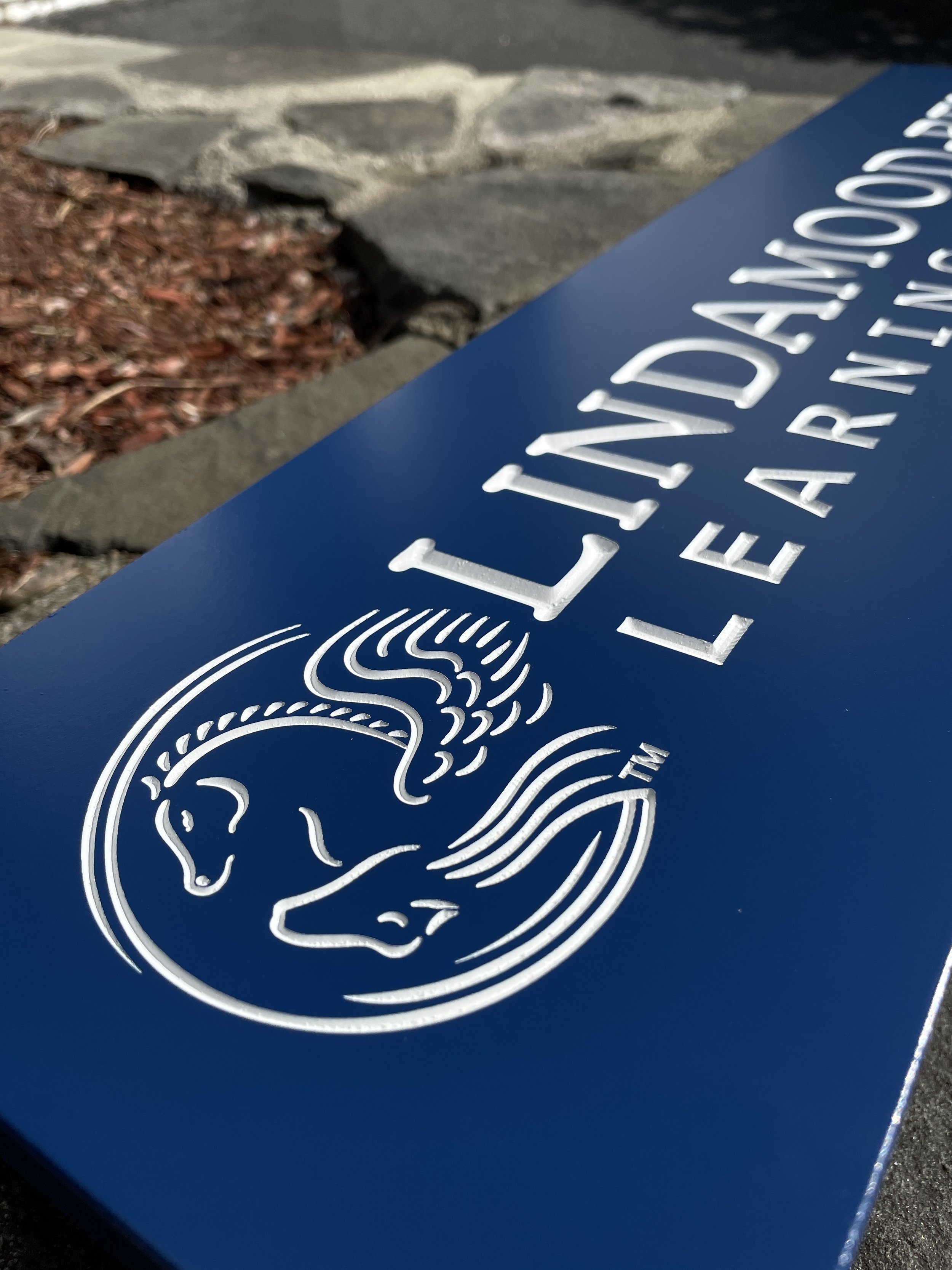 Close-up of a blue sign with the Lincoln Learning logo, featuring a stylized white line drawing of a horse's head and a doll's face, with the words "Lincoln Learning" printed above.