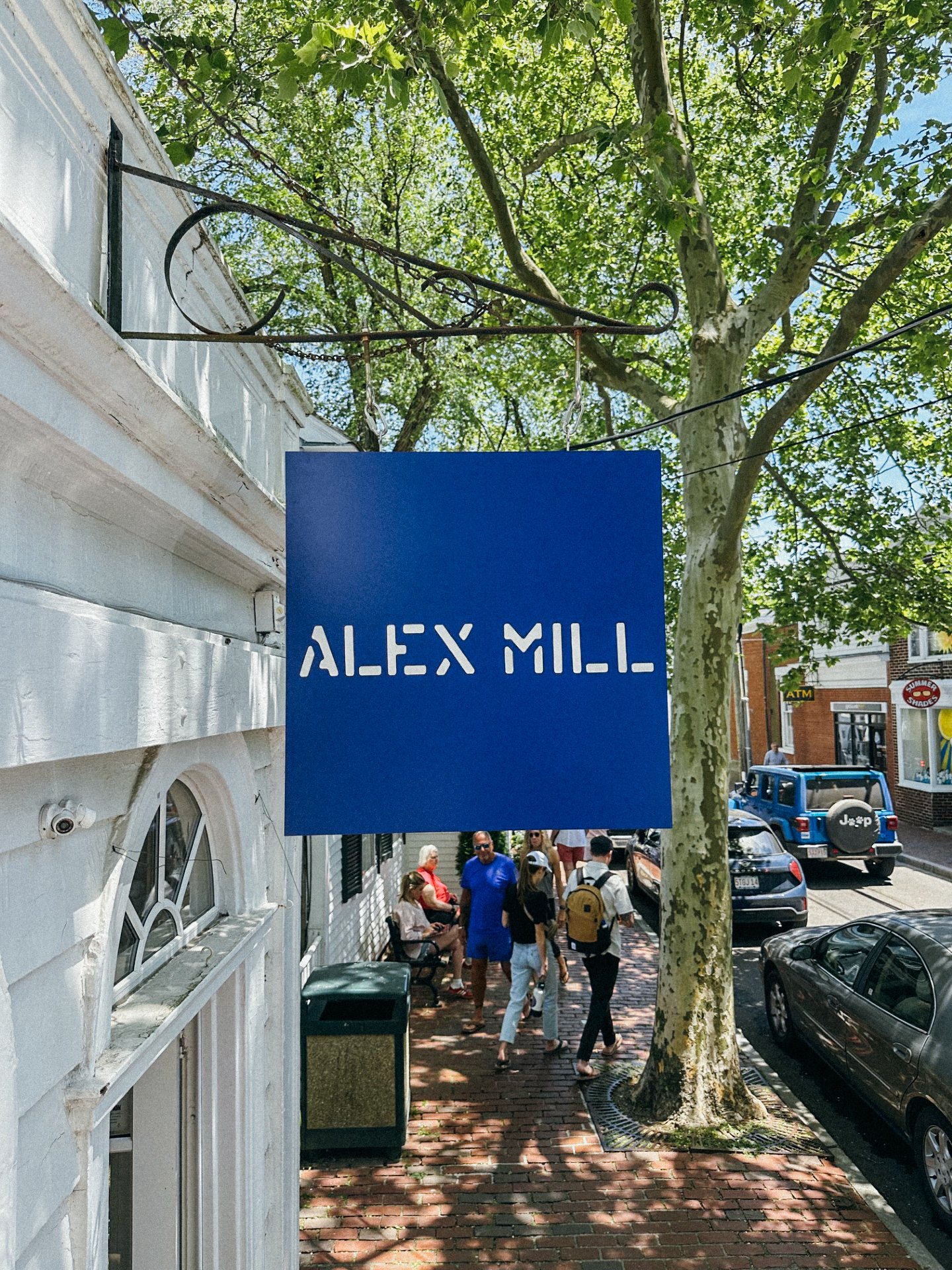 Blue sign with white text reading 'ALEX MILL' hanging on a street with pedestrians, parked cars, and trees.