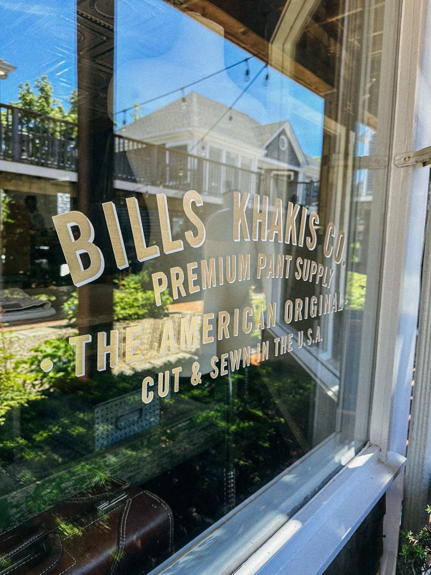 Shop window reflecting houses and trees outside, with a sign that reads 'BILLS KHAKIS CO. PREMIUM PANT SUPPLY THE AMERICAN ORIGINAL CUT & SEWN IN THE U.S.A.'
