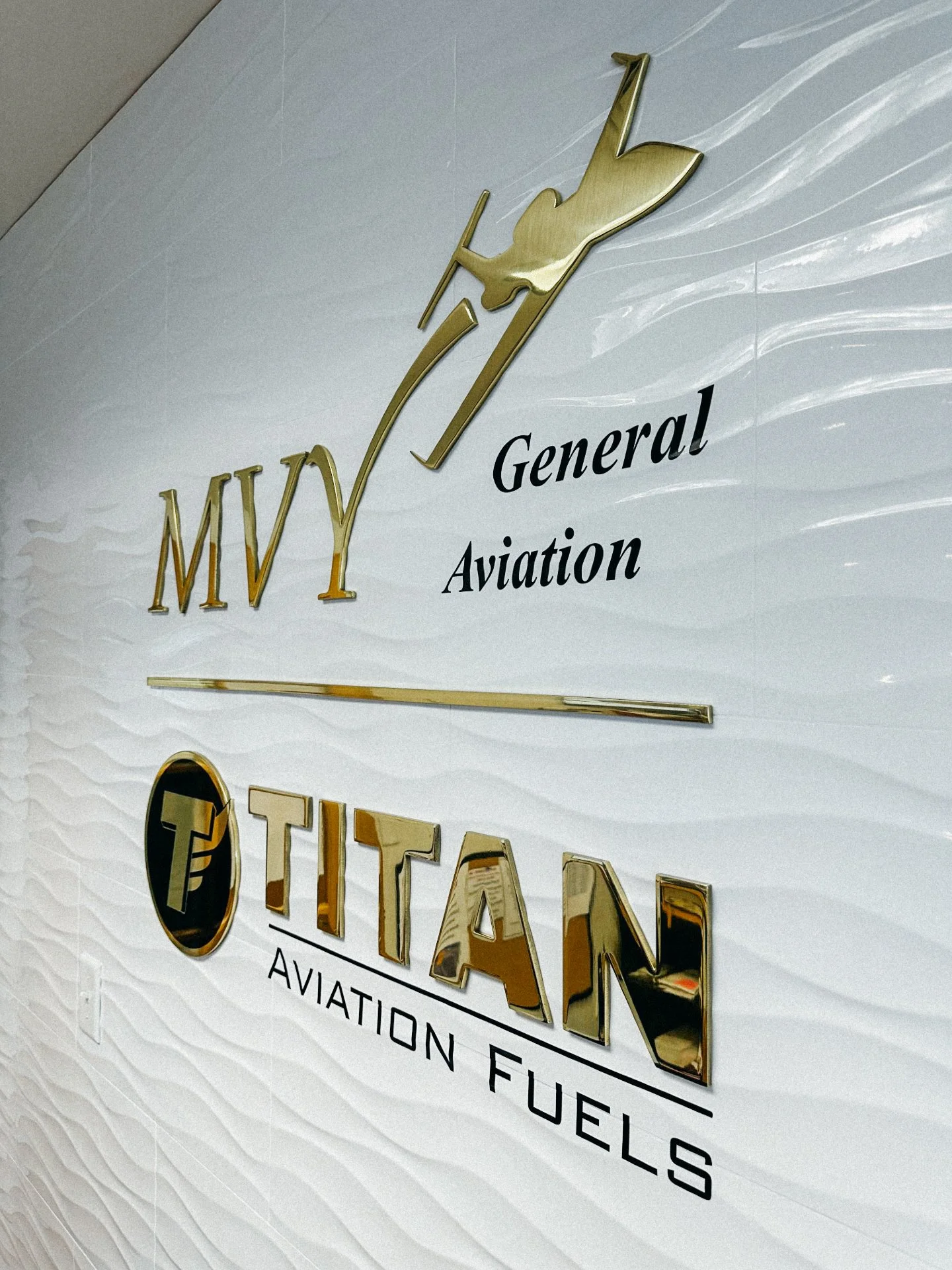 Sign for Moe General Aviation, featuring a metallic logo with a stylized aircraft and text in black and gold, on a textured white background.