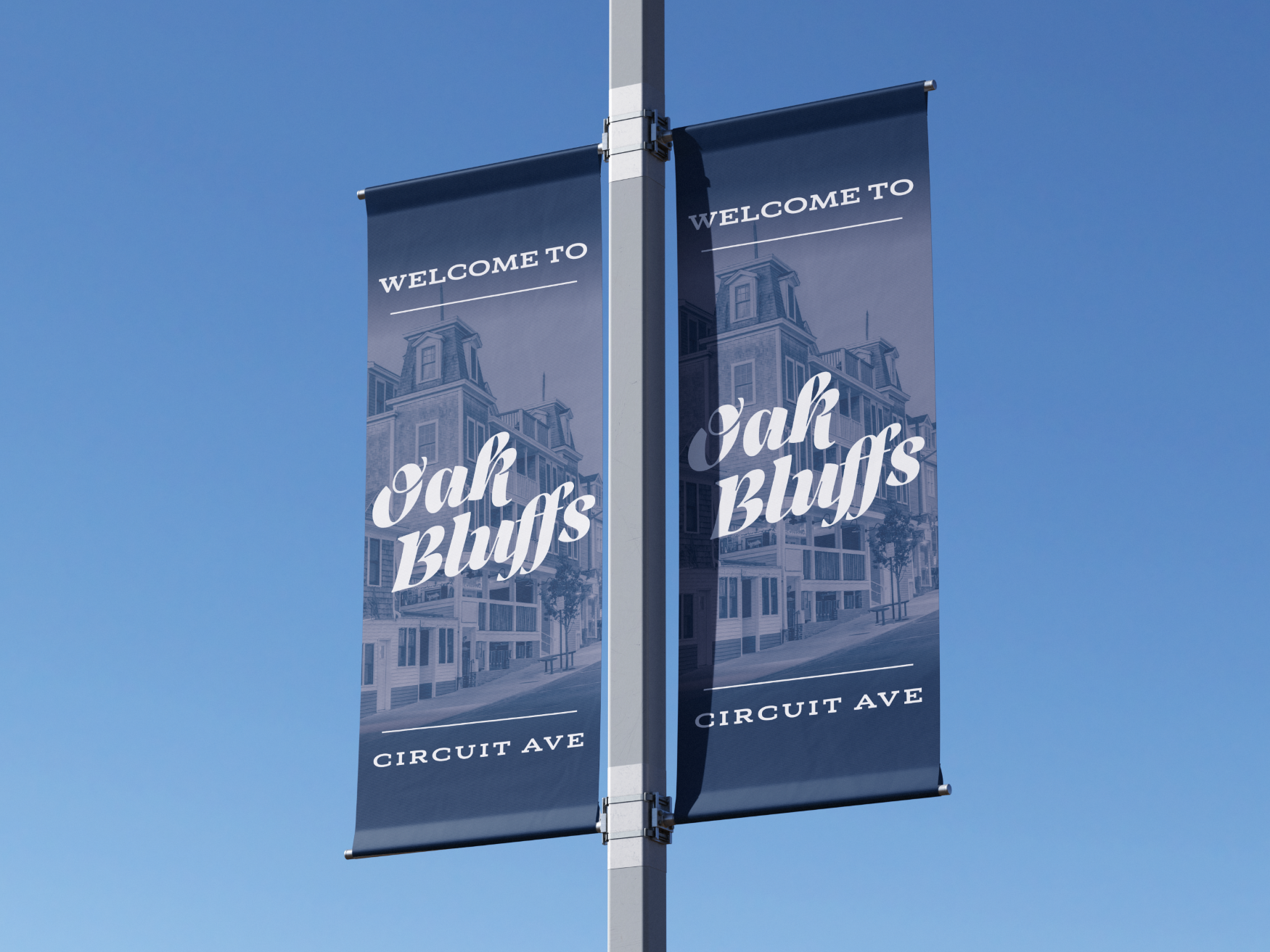 Two vertical banners on a street pole welcoming visitors to Oak Bluffs on Circuit Avenue, with an illustration of a Victorian-style house background, set against a clear blue sky.