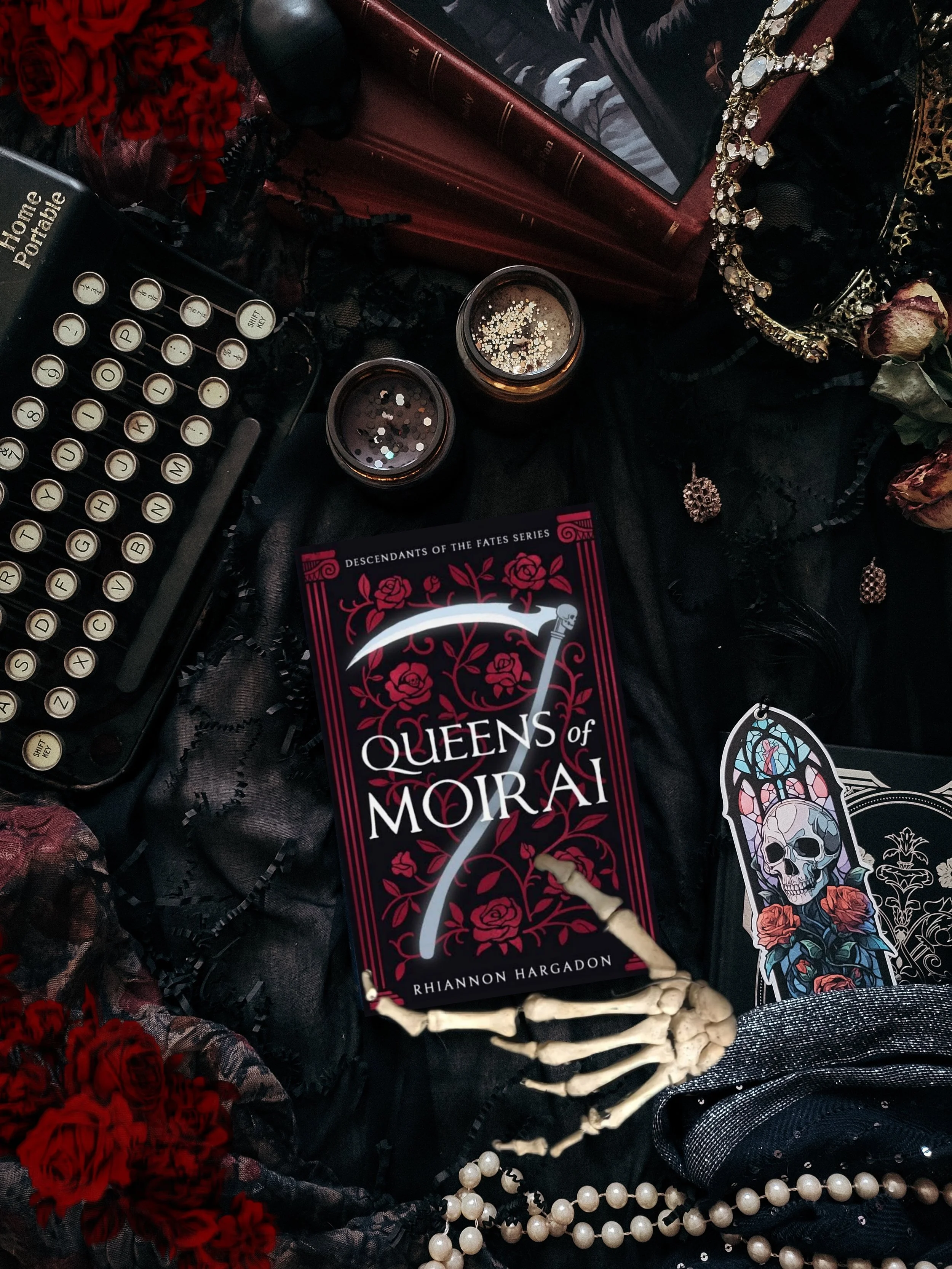A collection of gothic and dark-themed items includes a copy of the book 'Queens of Moral' by Riannon Hargadon, a skeleton hand, pearl necklaces, a skull sticker, a skull and roses sticker, two candles with dark glitter, a large red book, a photograp