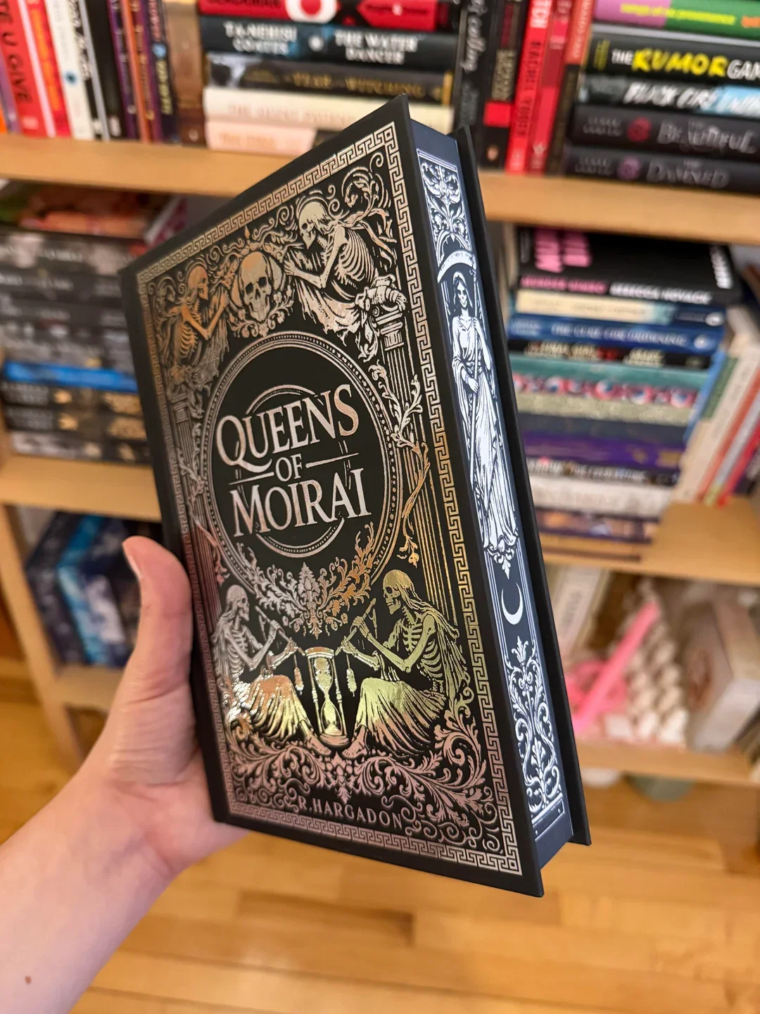 Book titled "Queens of Moira" with black and gold cover design featuring skeletons, skulls, and ornate patterns, held in a store with shelves of various books in the background.