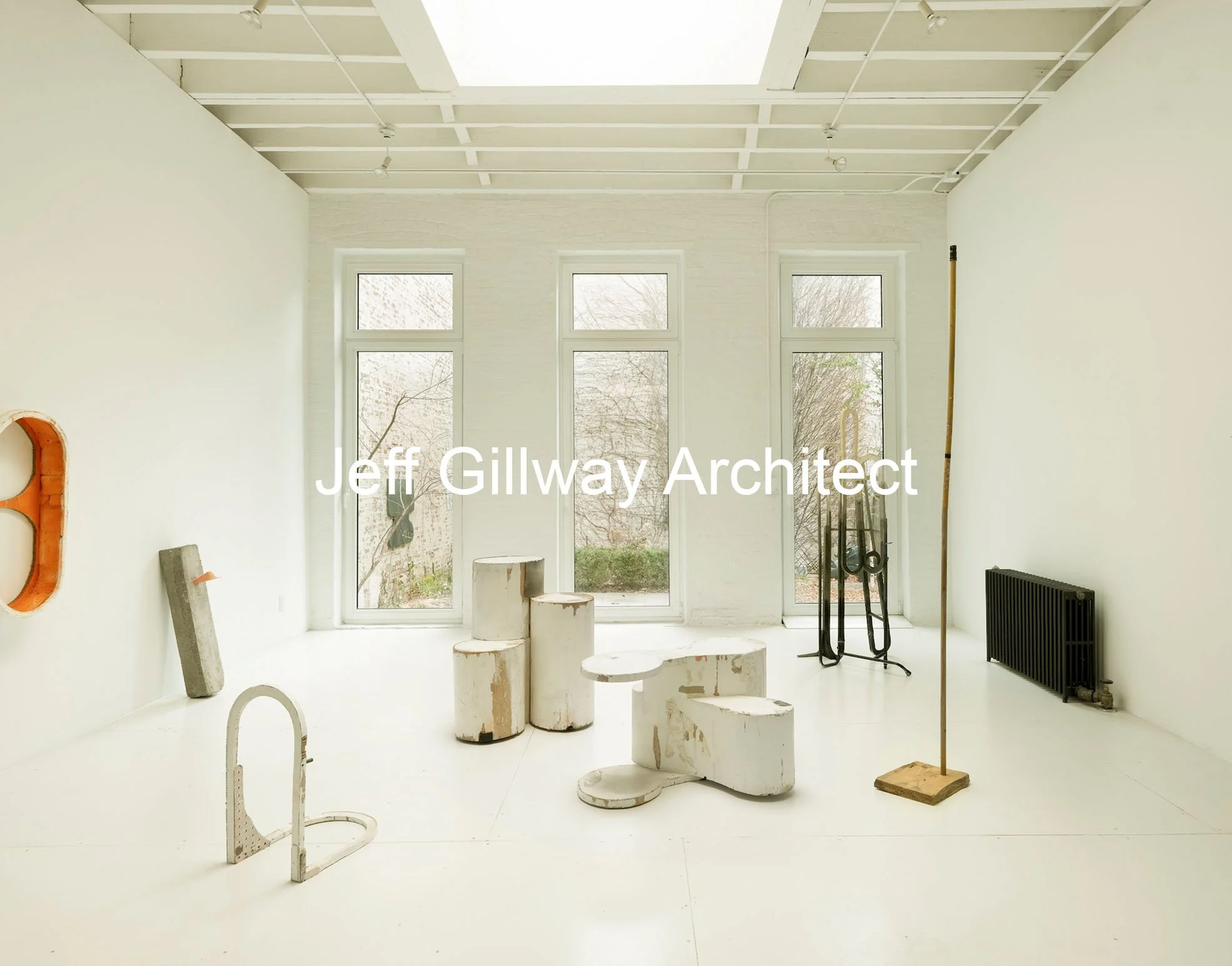 Minimalist art studio with white walls and large windows, featuring various sculptural objects, including cylindrical wooden sculptures, metal frames, and abstract art.