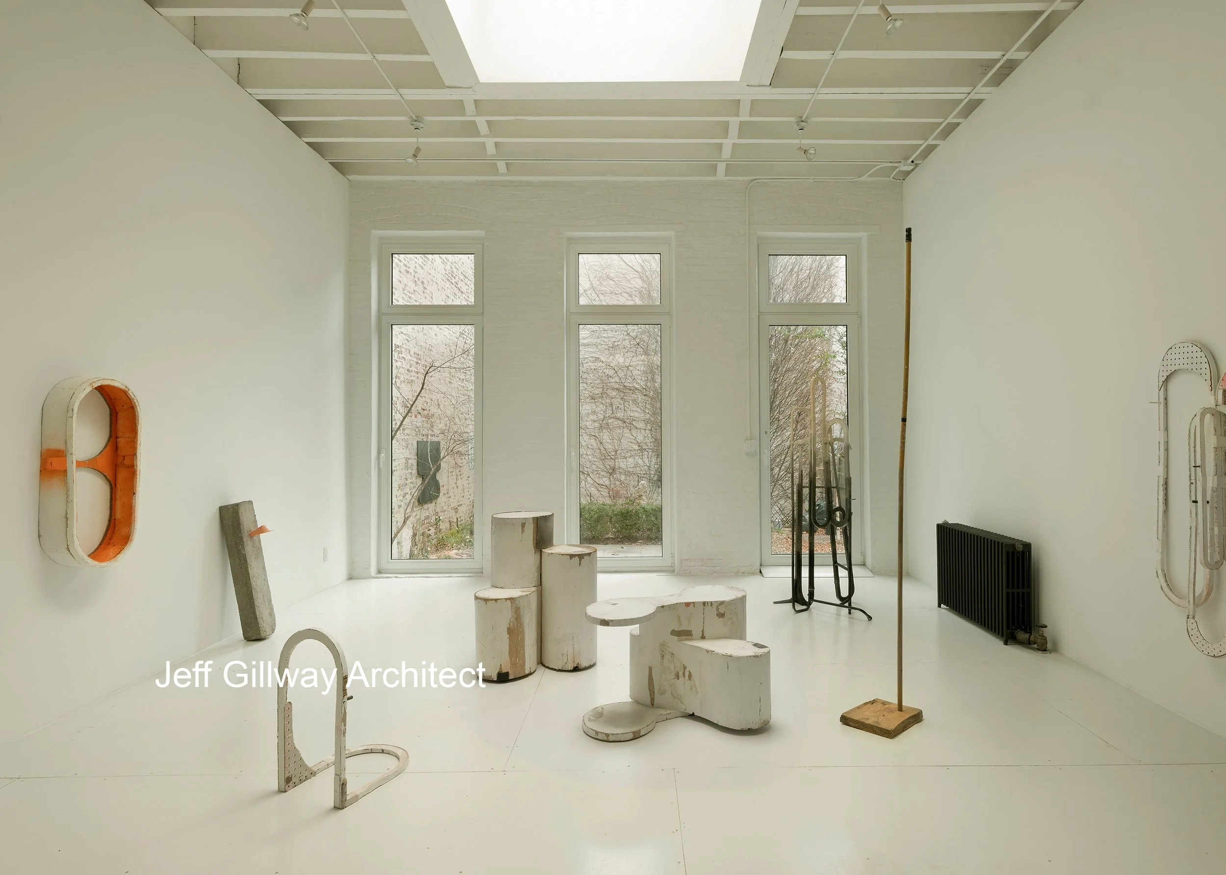Minimalist art installation in a gallery space with white walls and large windows, featuring abstract sculptural objects, a black radiator, and modern art.