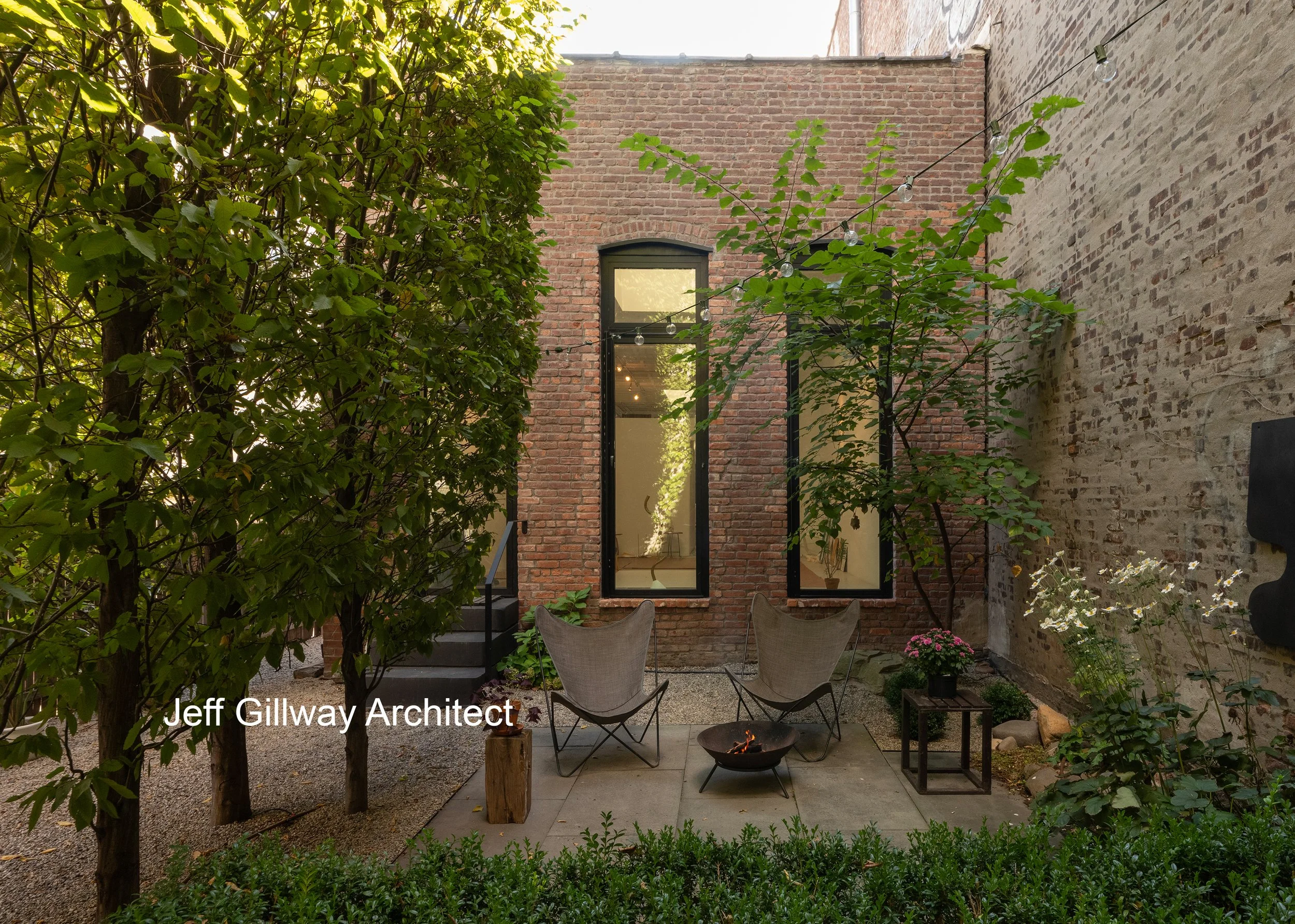 A cozy outdoor patio area with two chairs, a small fire pit, and lush green plants surrounding a brick building with large windows.