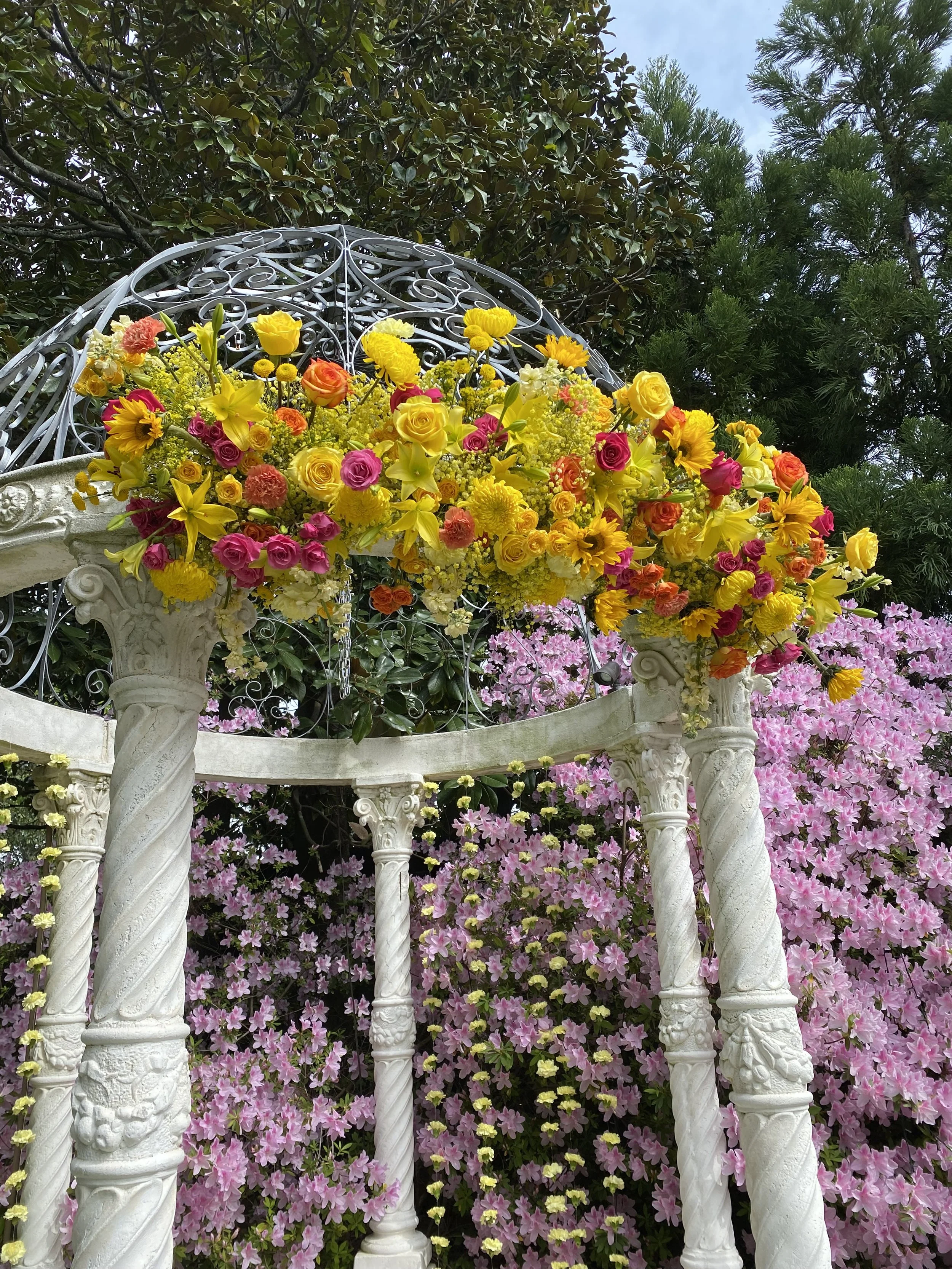 A decorative garden arch adorned with vibrant yellow, pink, and orange flowers, set amidst pink flowering bushes and green trees.