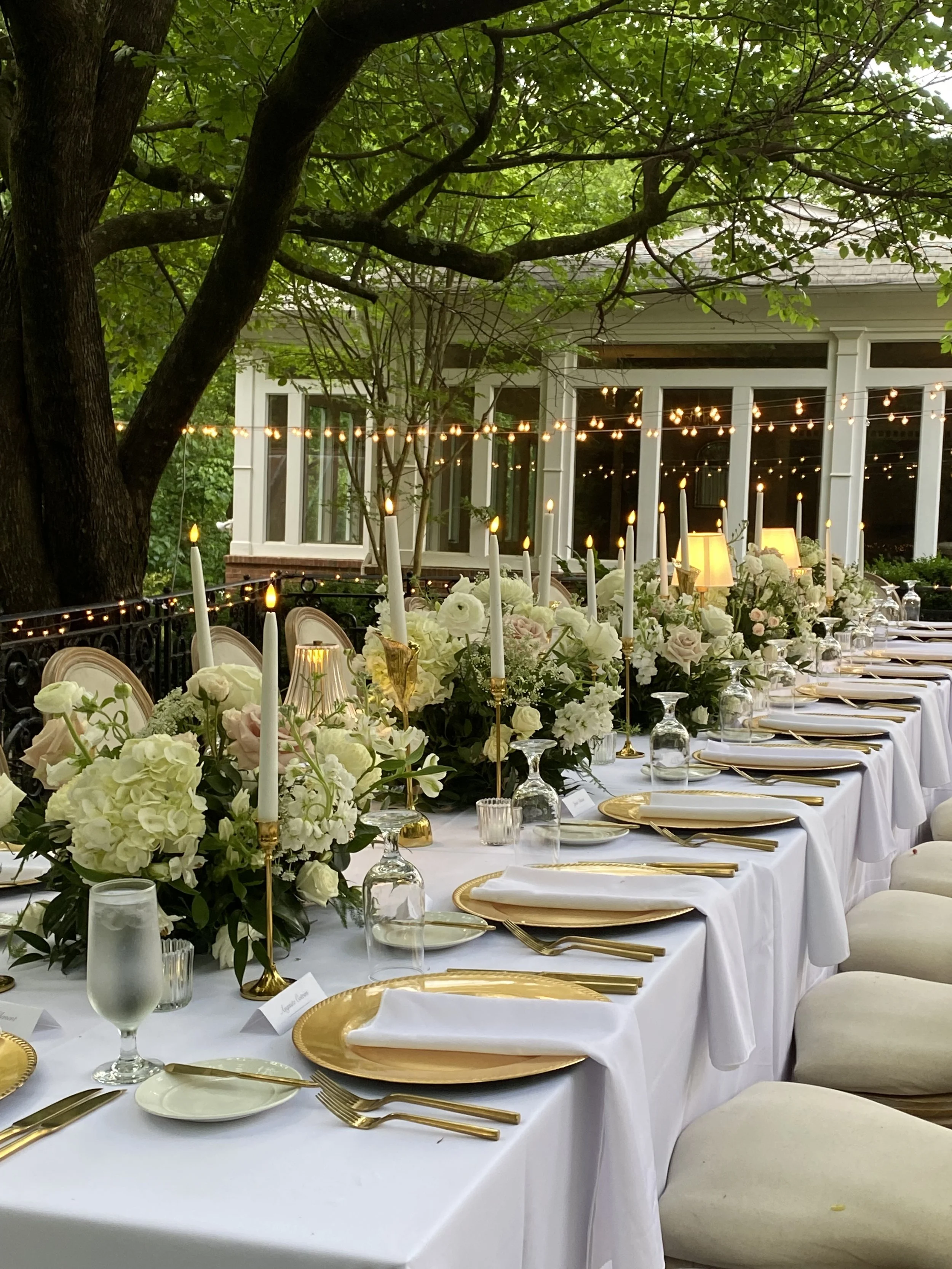 Elegant outdoor dining table set with white linens, gold chargers, white napkins, gold cutlery, tall white candles, floral centerpieces, and string lights under a large leafy tree.