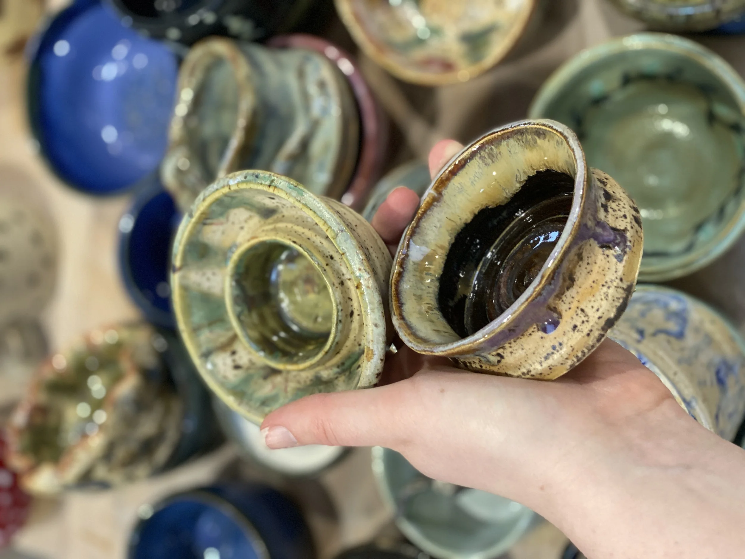 A hand holding a small glazed ceramic bowl or cup, with several more glazed bowls or cups in different colors and glazes arranged in the background.