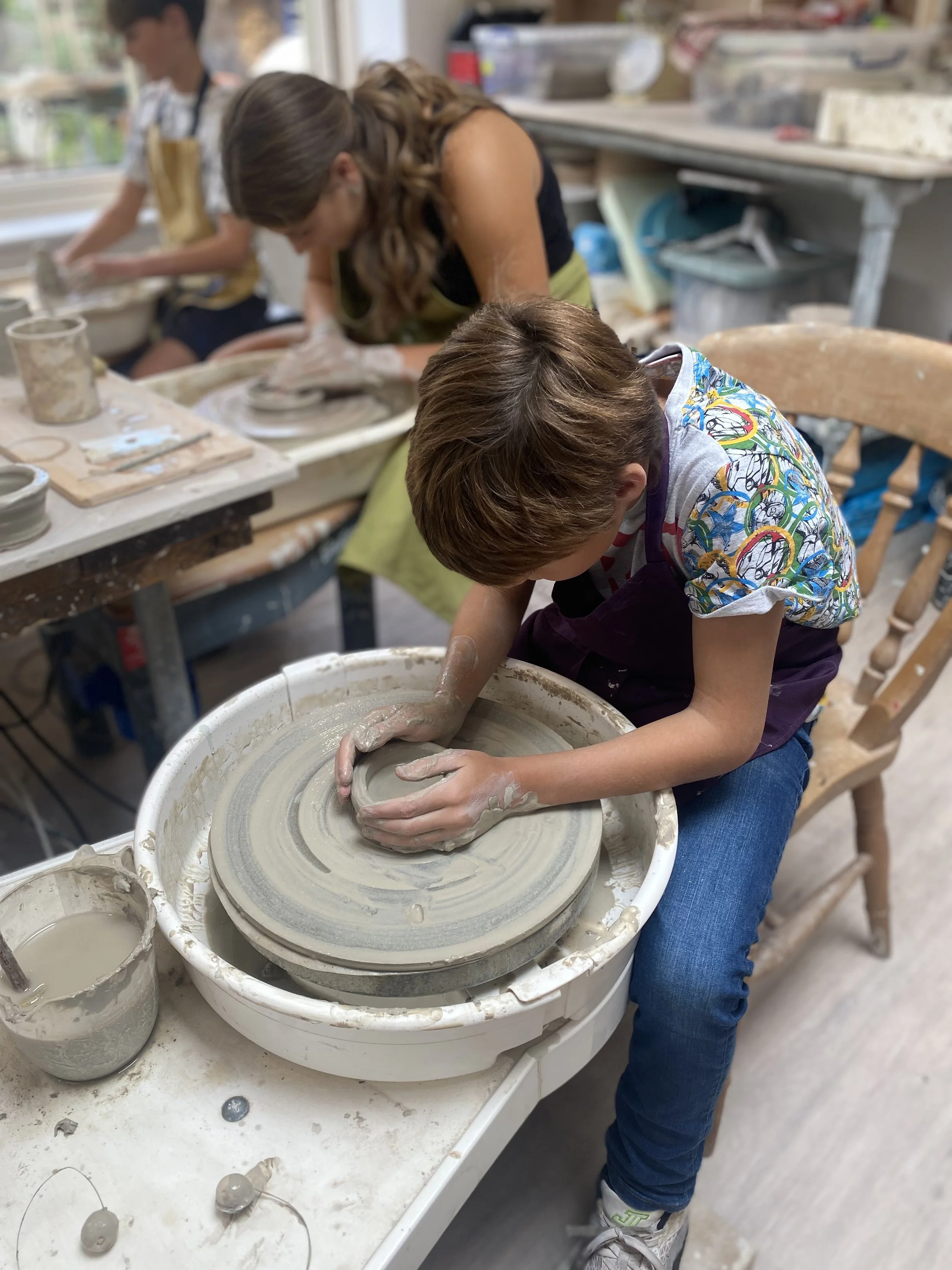 A boy is sitting on a wooden chair, shaping clay on a pottery wheel in a pottery studio. Two other people, a woman and a girl, are working at a nearby work table with pottery tools and supplies.