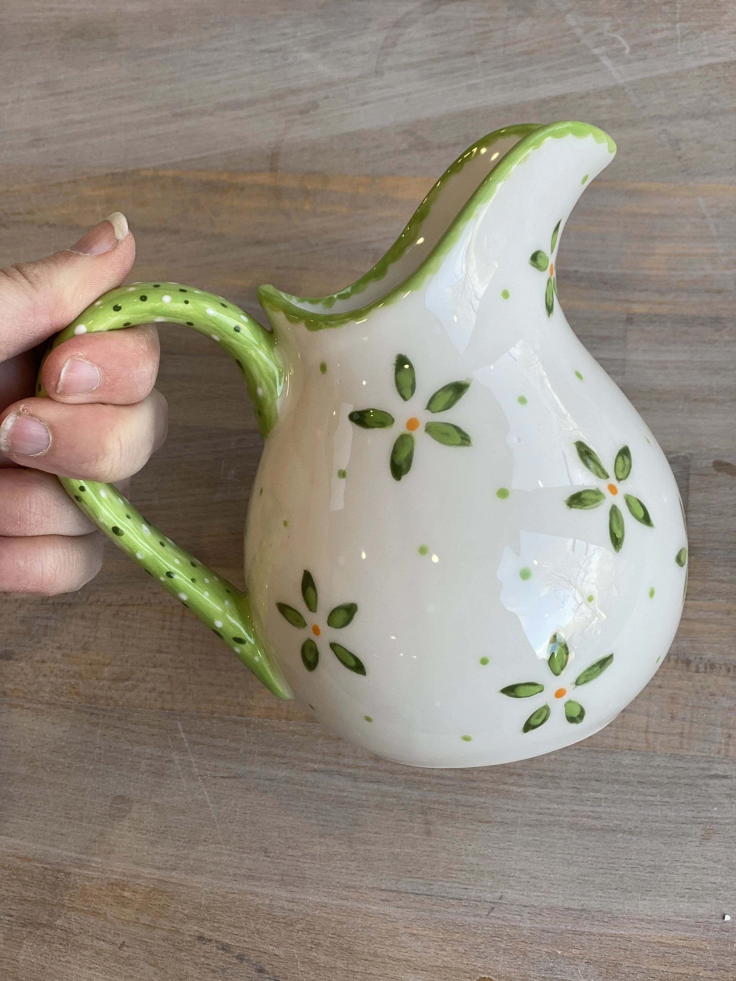 A ceramic pitcher with a white body and green floral designs, featuring a green polka-dotted handle and rim, placed on a wooden surface.