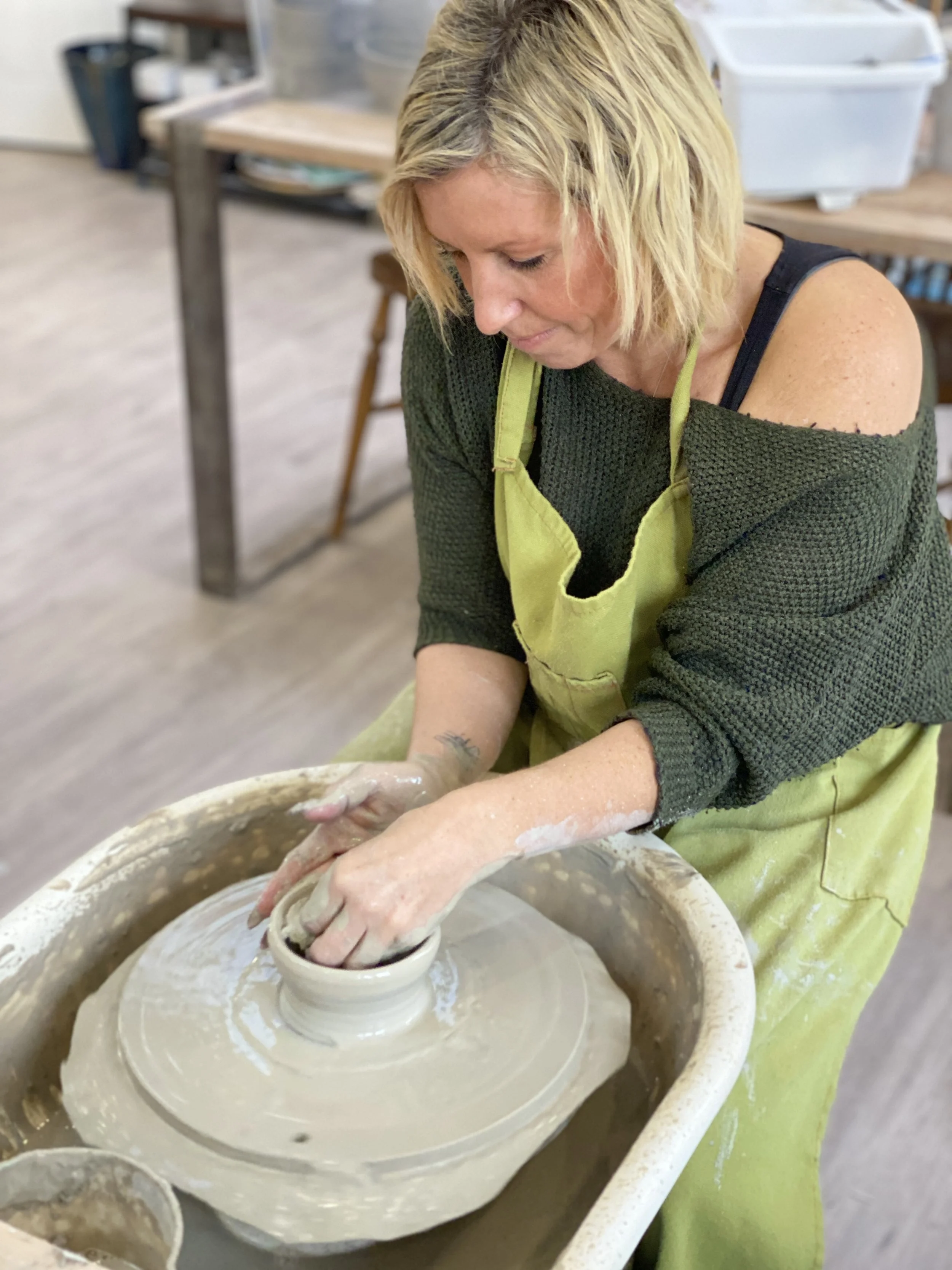 Woman with blonde hair practicing pottery, shaping clay on a potter's wheel in a studio.