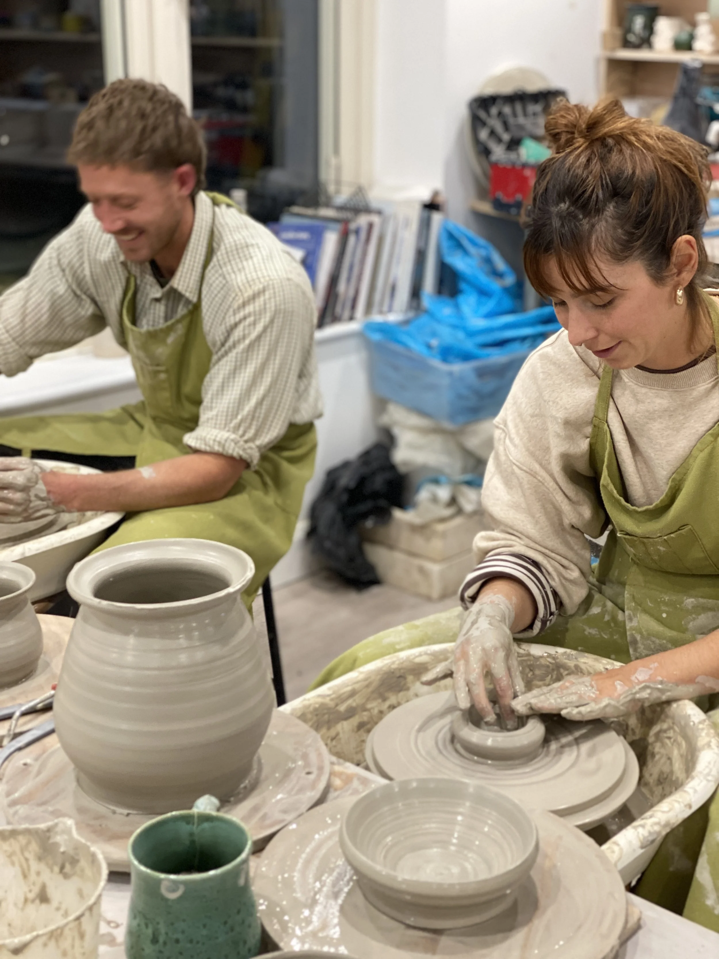 A man and woman are working on pottery in a ceramics studio, both wearing green aprons and covered in clay.