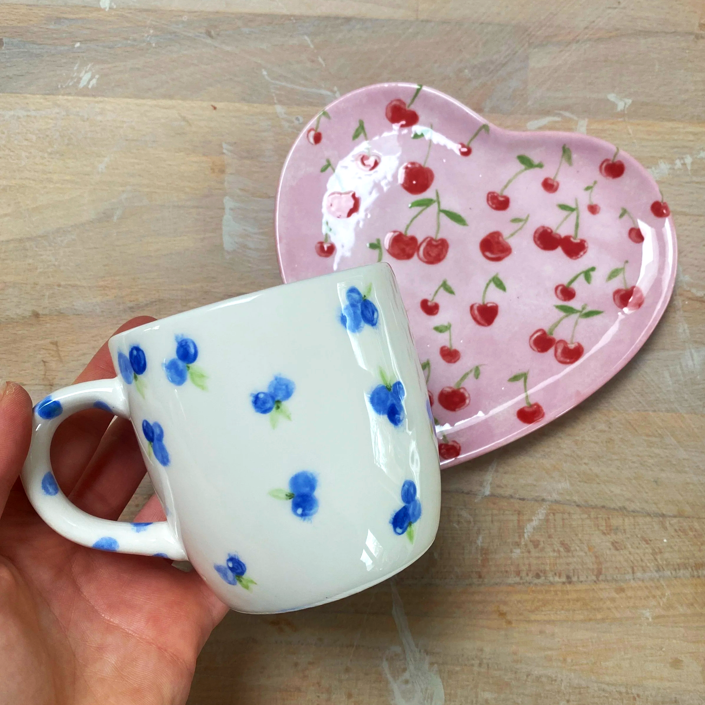 A white ceramic mug with blue floral patterns held by a hand, with a pink heart-shaped dish decorated with cherries in the background on a wooden surface.