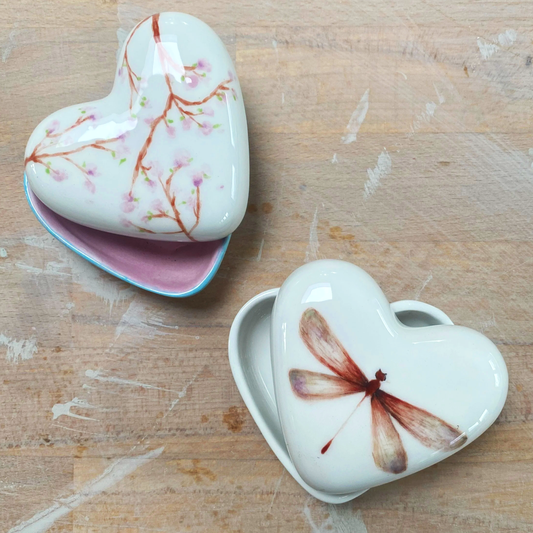 Two heart-shaped ceramic boxes with floral and dragonfly designs on a wooden surface.