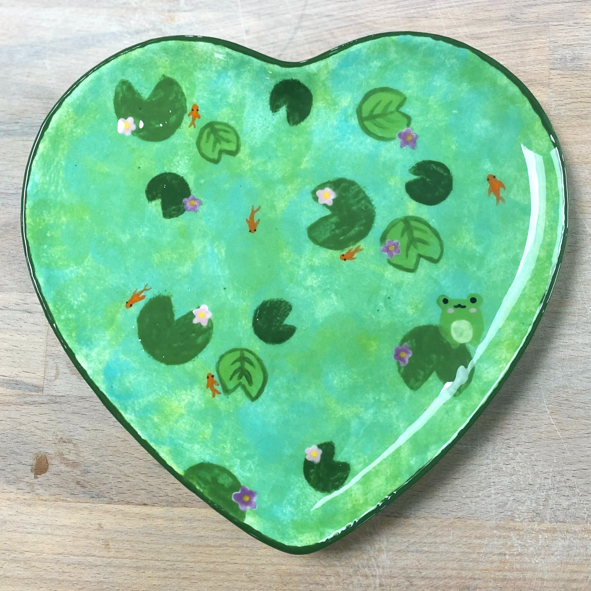 Decorative heart-shaped dish with painted lily pads, small frogs, fish, and purple flowers in shades of green, purple, orange, and white.