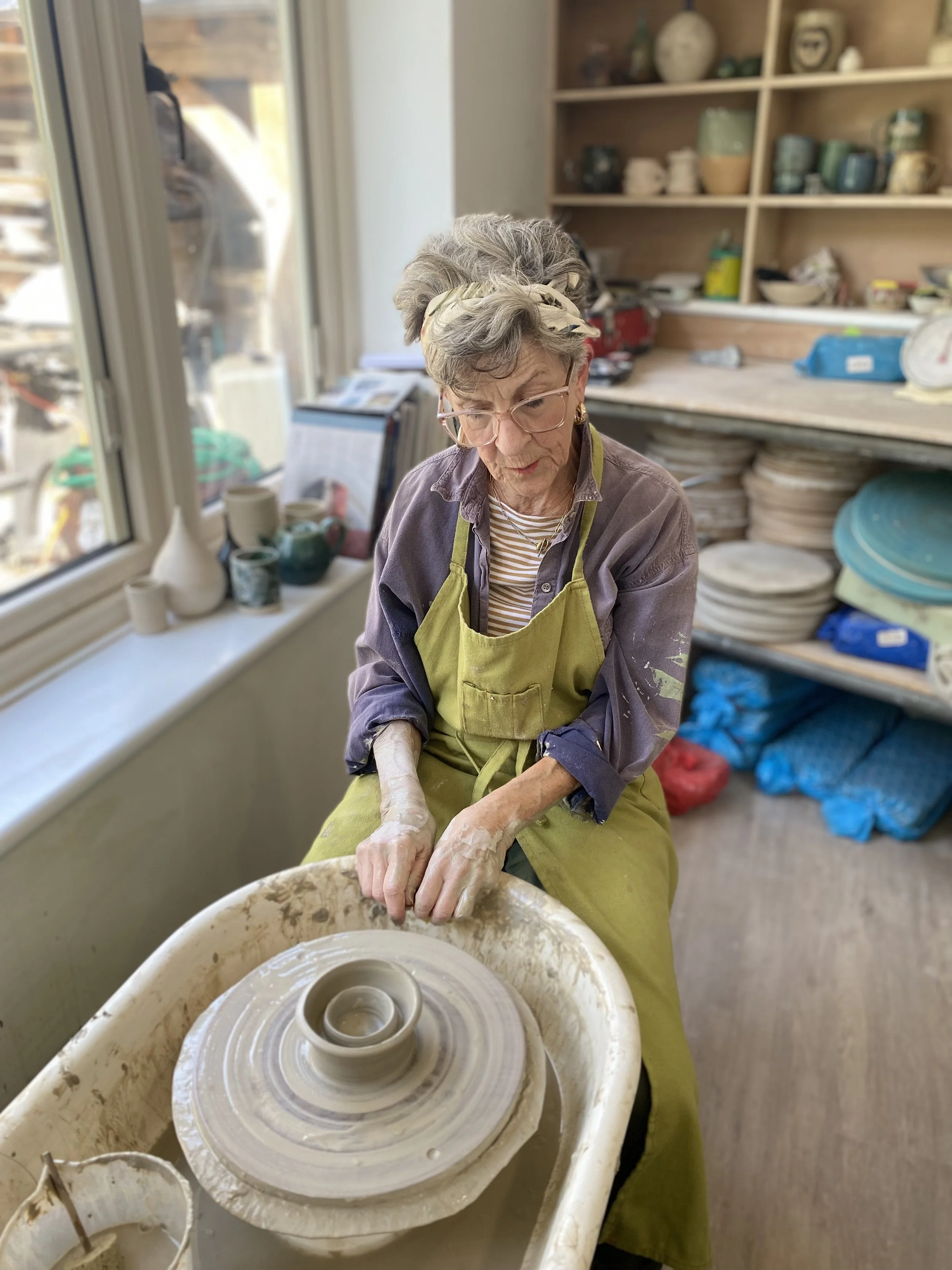 An elderly woman with glasses, wearing a headband and a purple shirt under a green apron, is sitting at a pottery wheel shaping a piece of clay into a small vase or cup.
