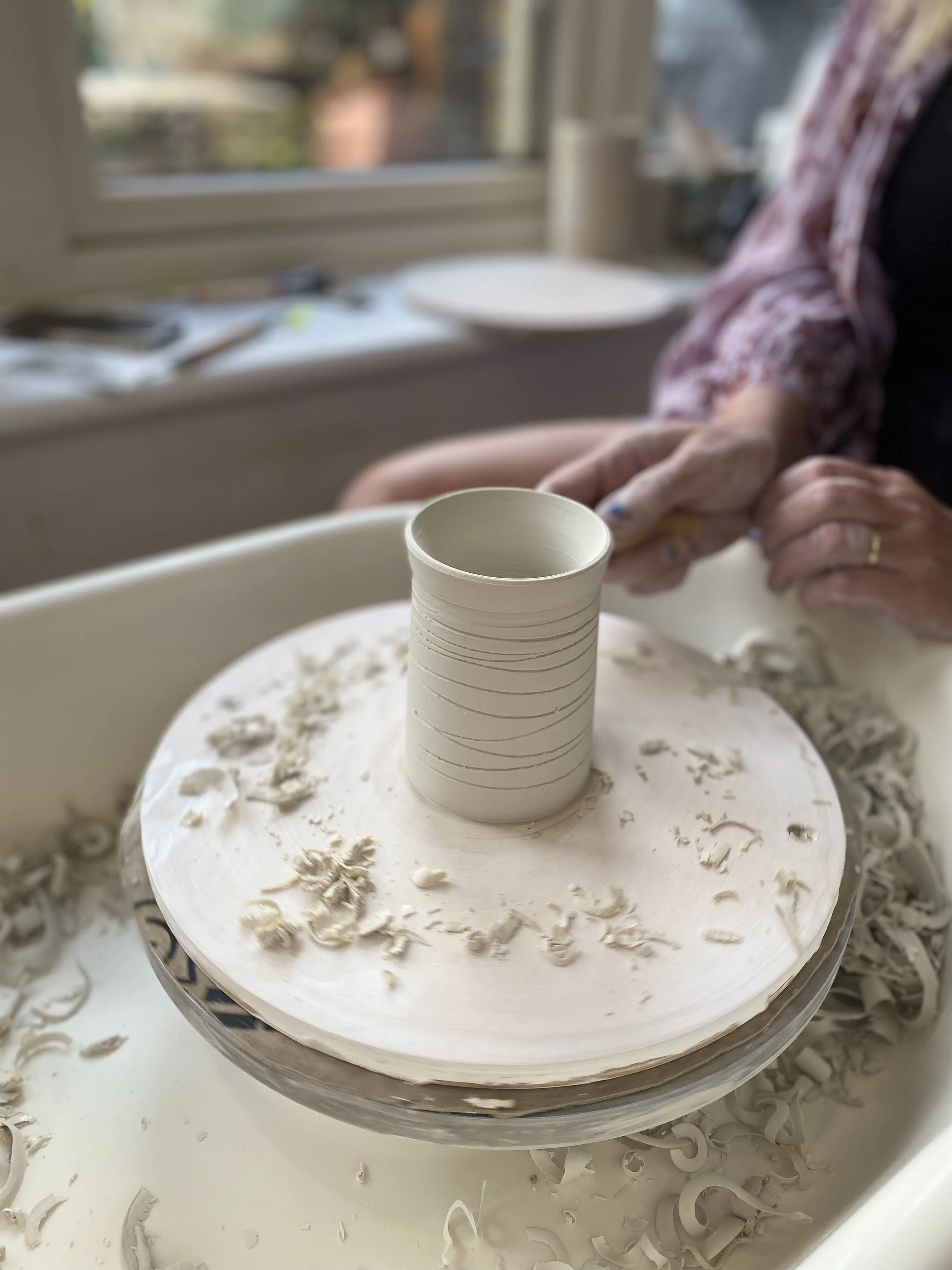 Close-up of a ceramic cup on a pottery wheel with a person shaping clay in the background.