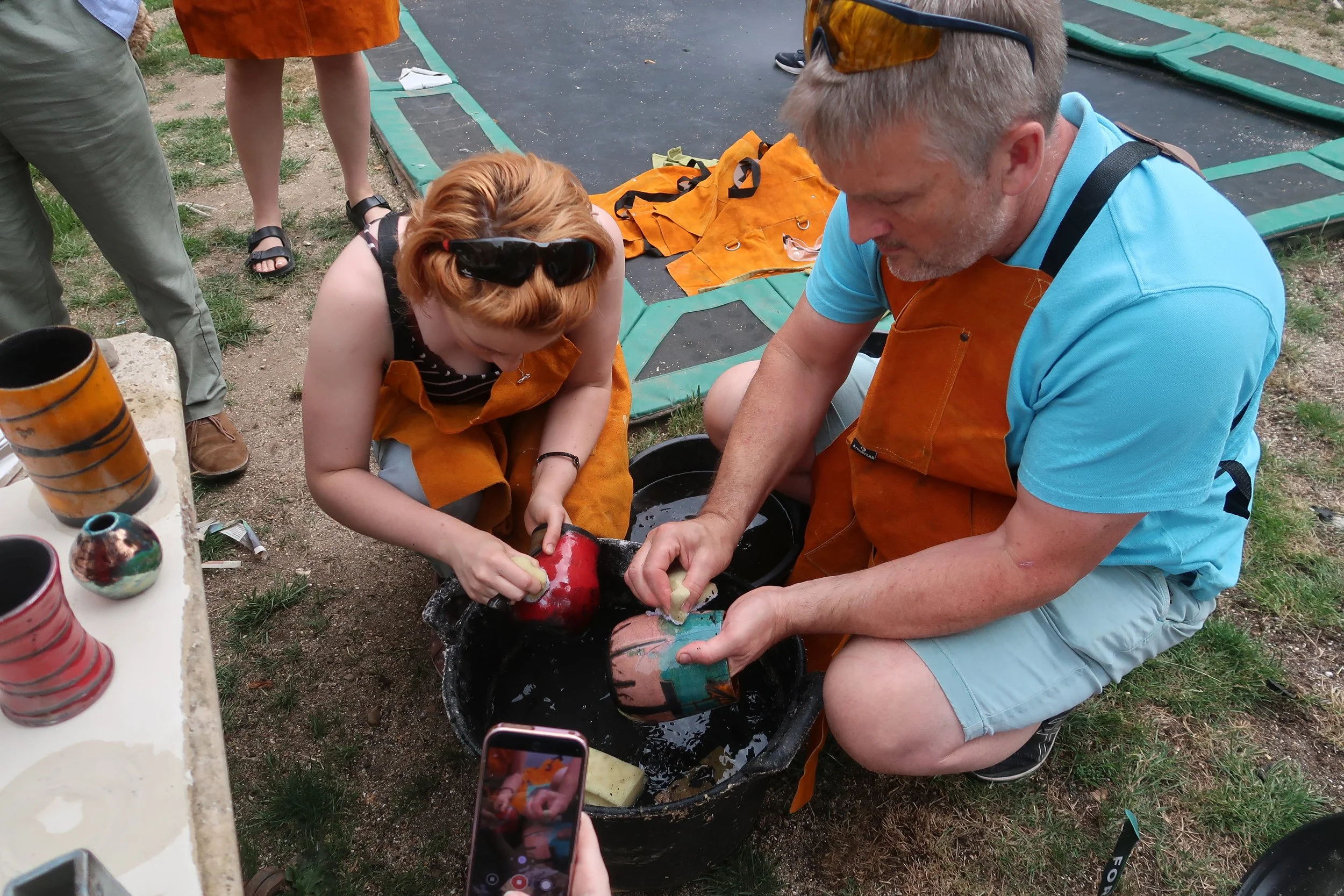 Two people, a woman with glasses on her head and a man, are involved in raku activities outdoors. The woman is holding a sponge claning pottery, and the man is holding a piece of colored ceramics over a water basin.