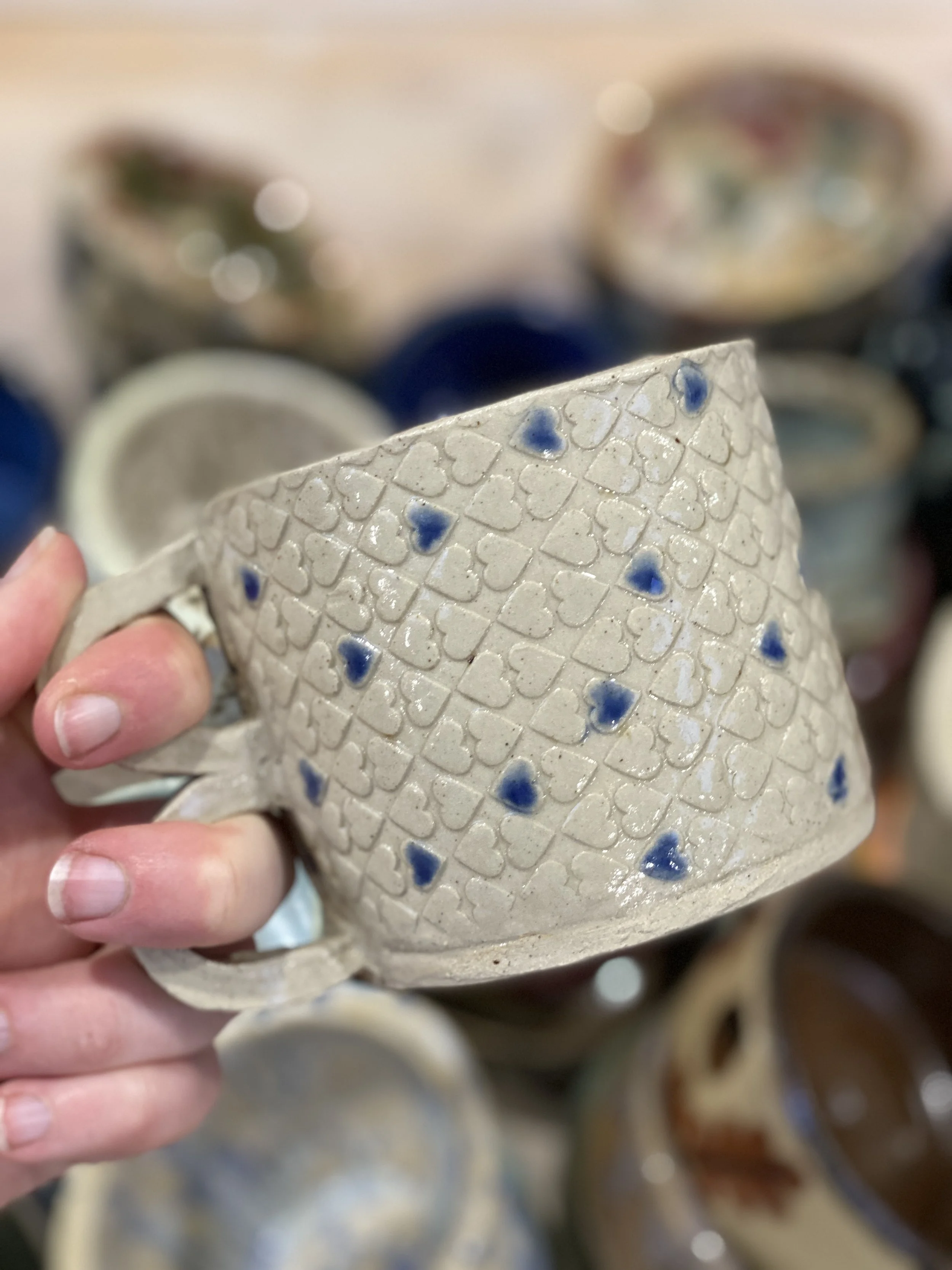 Person holding a ceramic mug decorated with blue hearts and grid patterns, with other similar mugs and a lemon slice in the background.