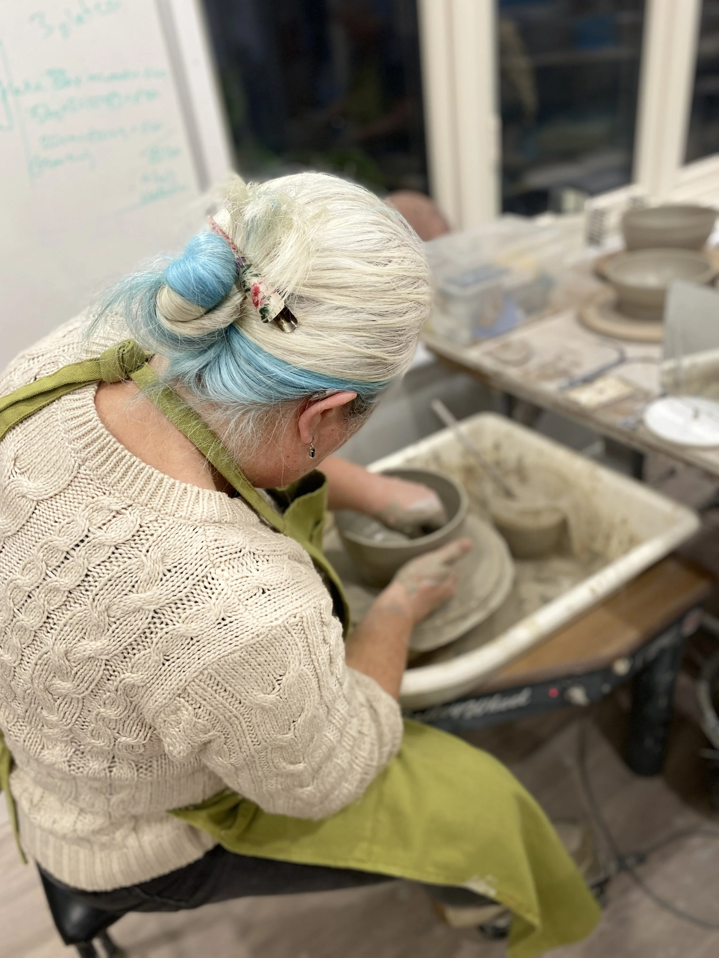 A woman with white and blue hair, wearing a beige knitted sweater and green apron, is shaping a clay pot at a pottery wheel in a pottery workshop.