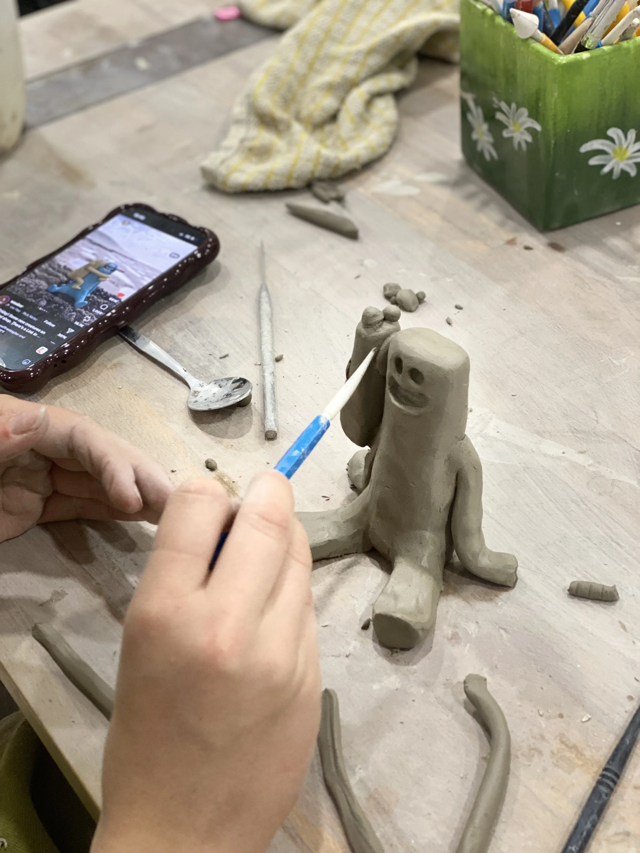 Child sculpting a clay figure with a smiling face on a tabletop, surrounded by sculpting tools, a phone with a social media app, and a container of colored pencils.