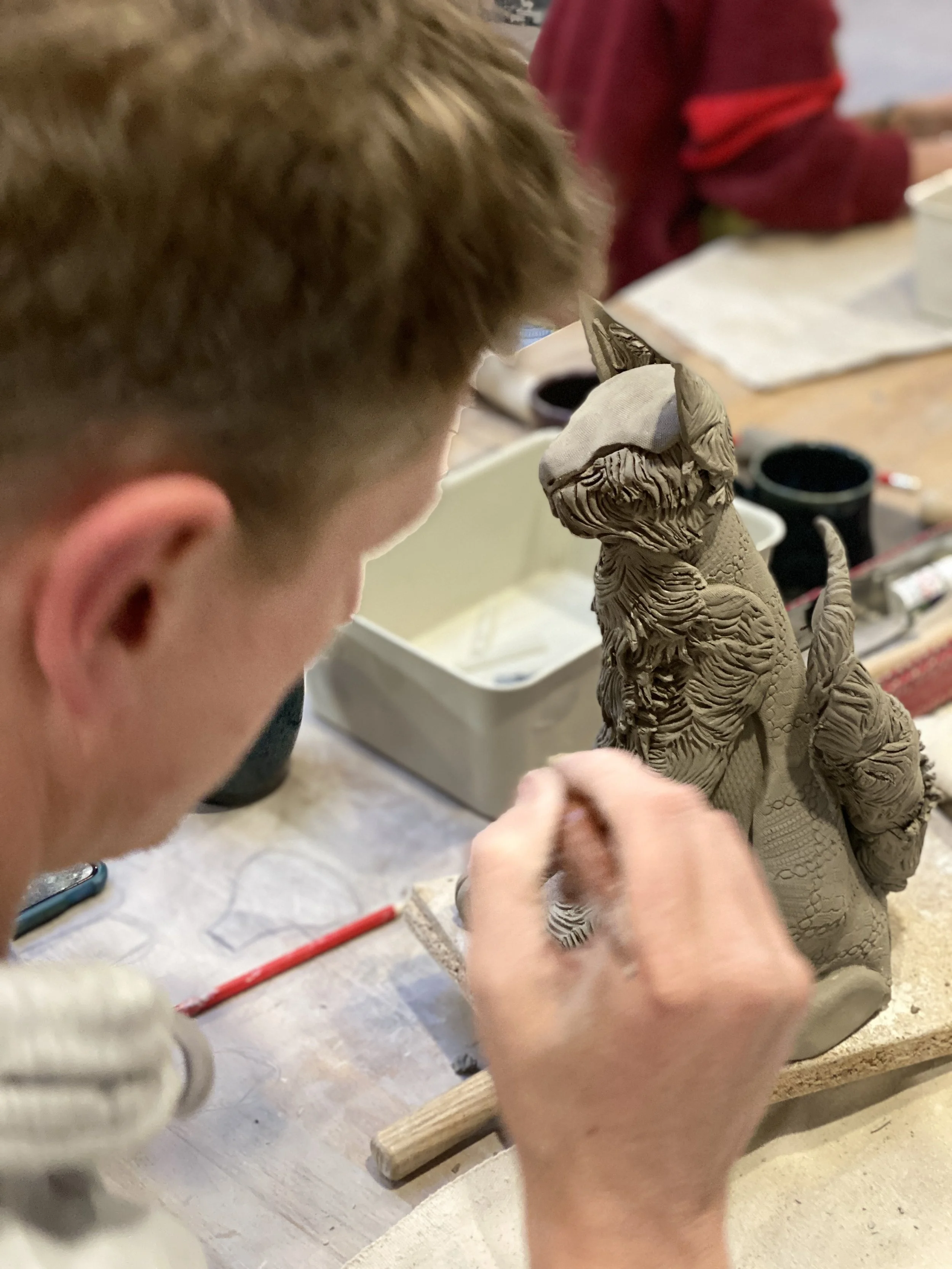 A person sculpting a detailed clay statue of a dog with a hat on. The workspace has various tools and supplies for sculpting.