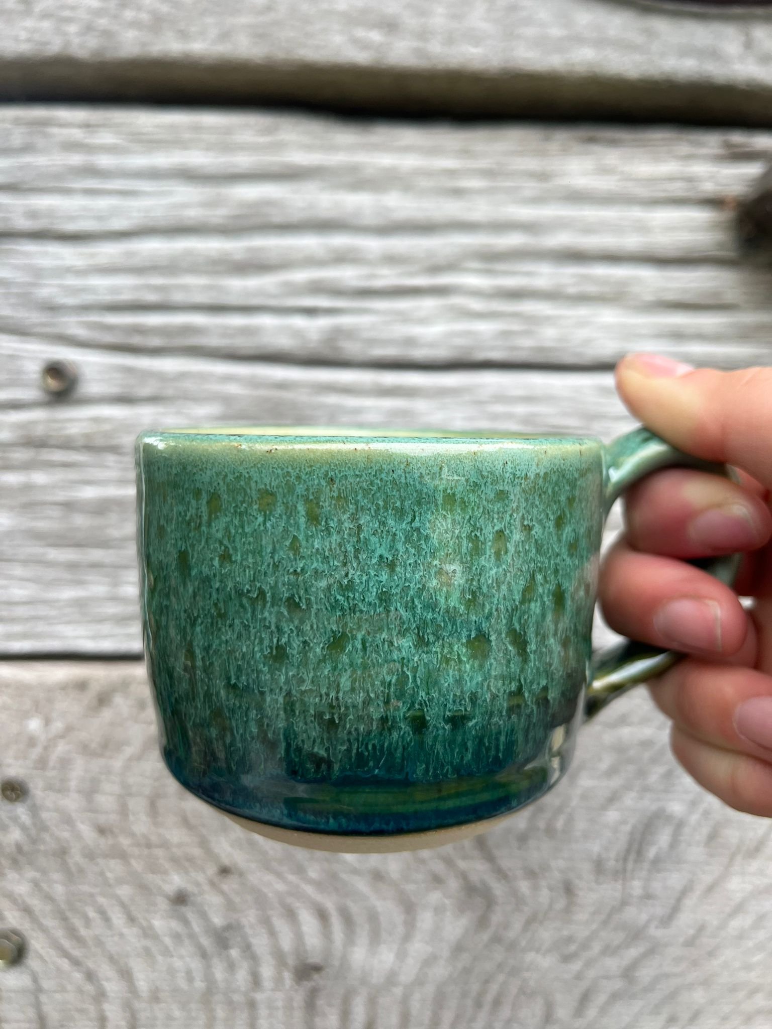 Hand holding a ceramic mug with a green and blue glaze in front of a weathered wooden surface