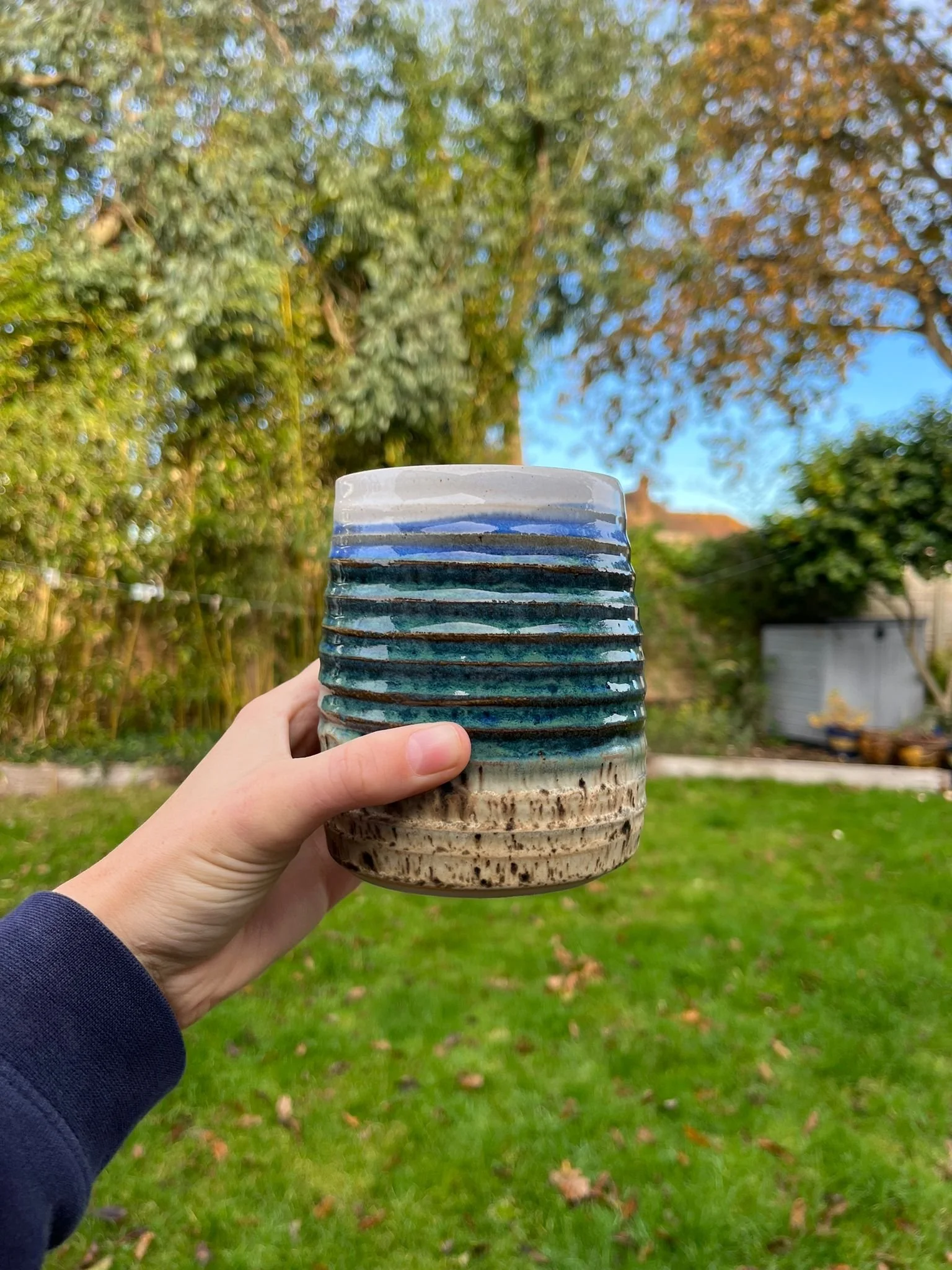 Person holding a colorful ceramic mug outdoors on a grassy lawn with trees and a shed in the background.