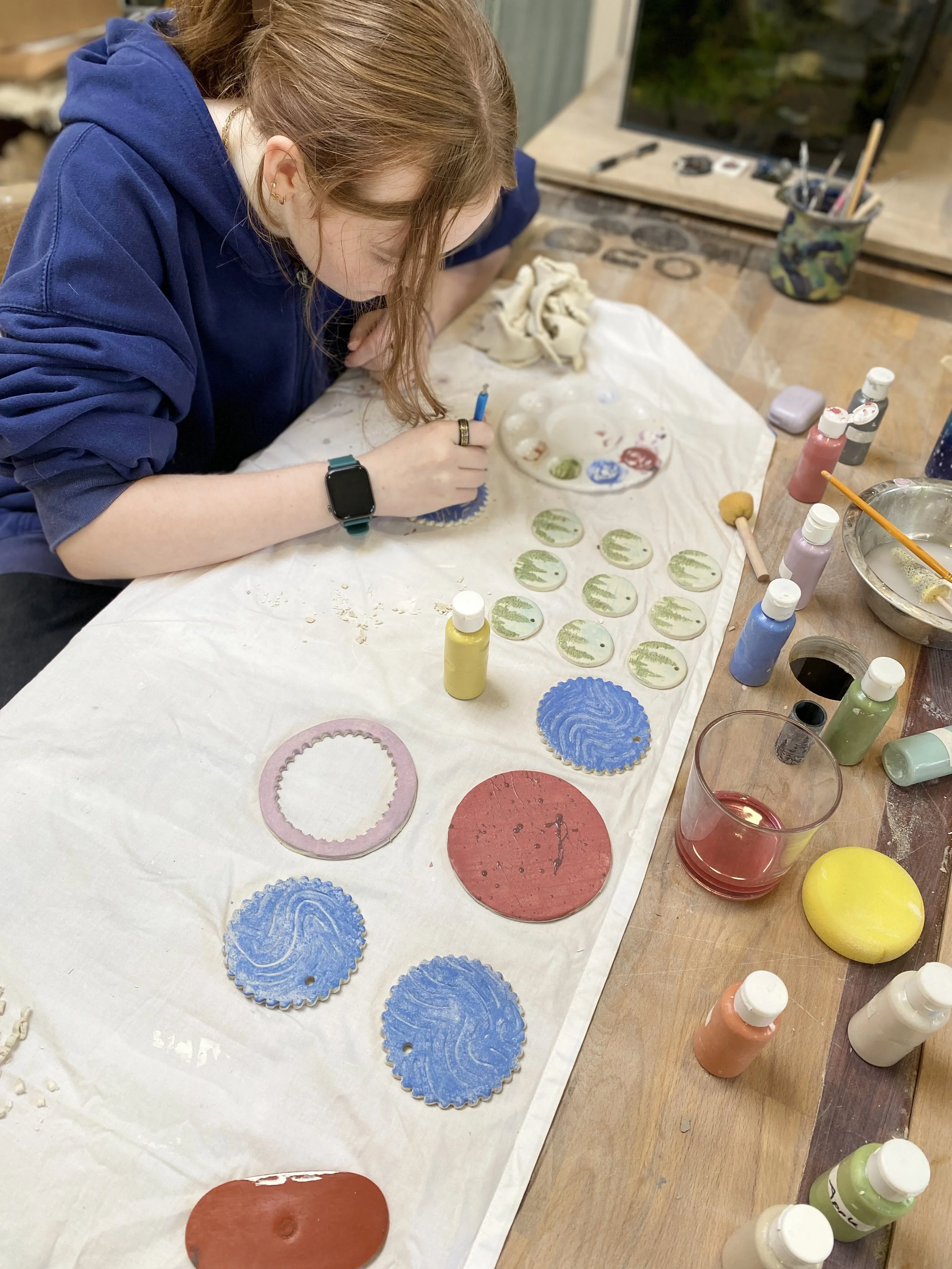 A person is painting circular ceramic tiles with green and blue designs on a worktable, surrounded by bottles of paint, brushes, and pottery tools.