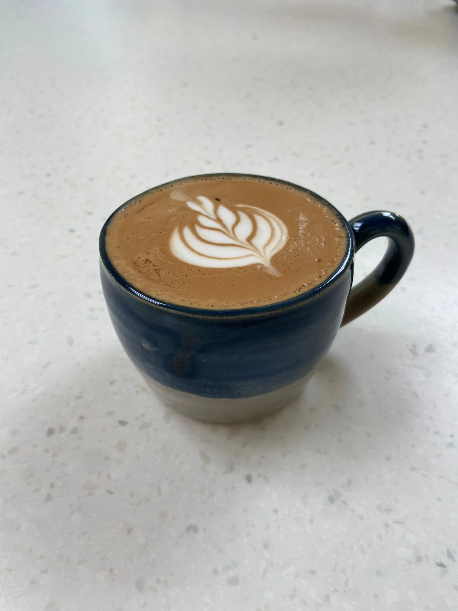 A blue ceramic mug filled with coffee topped with latte art on a white speckled countertop.