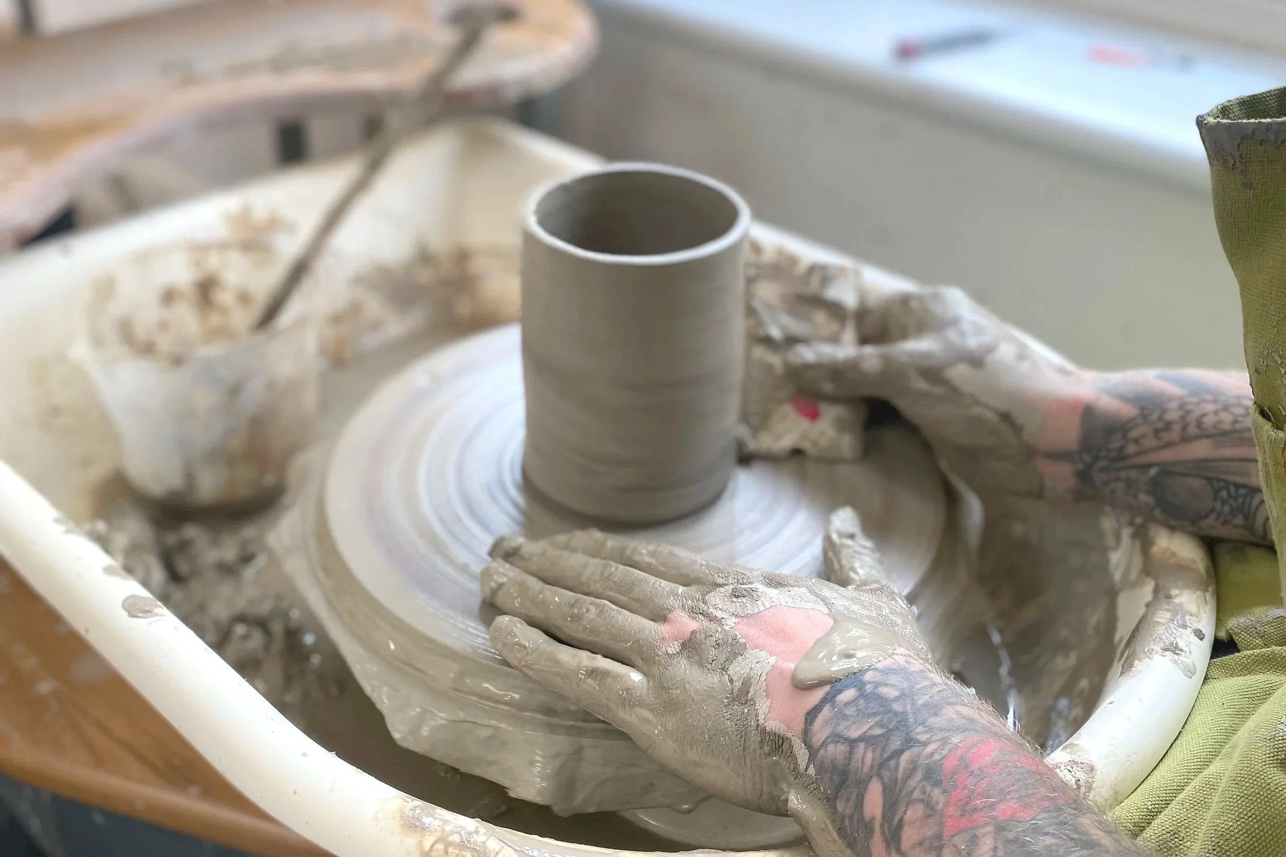 A person with tattooed hands shaping a ceramic mug on a pottery wheel in a pottery studio.