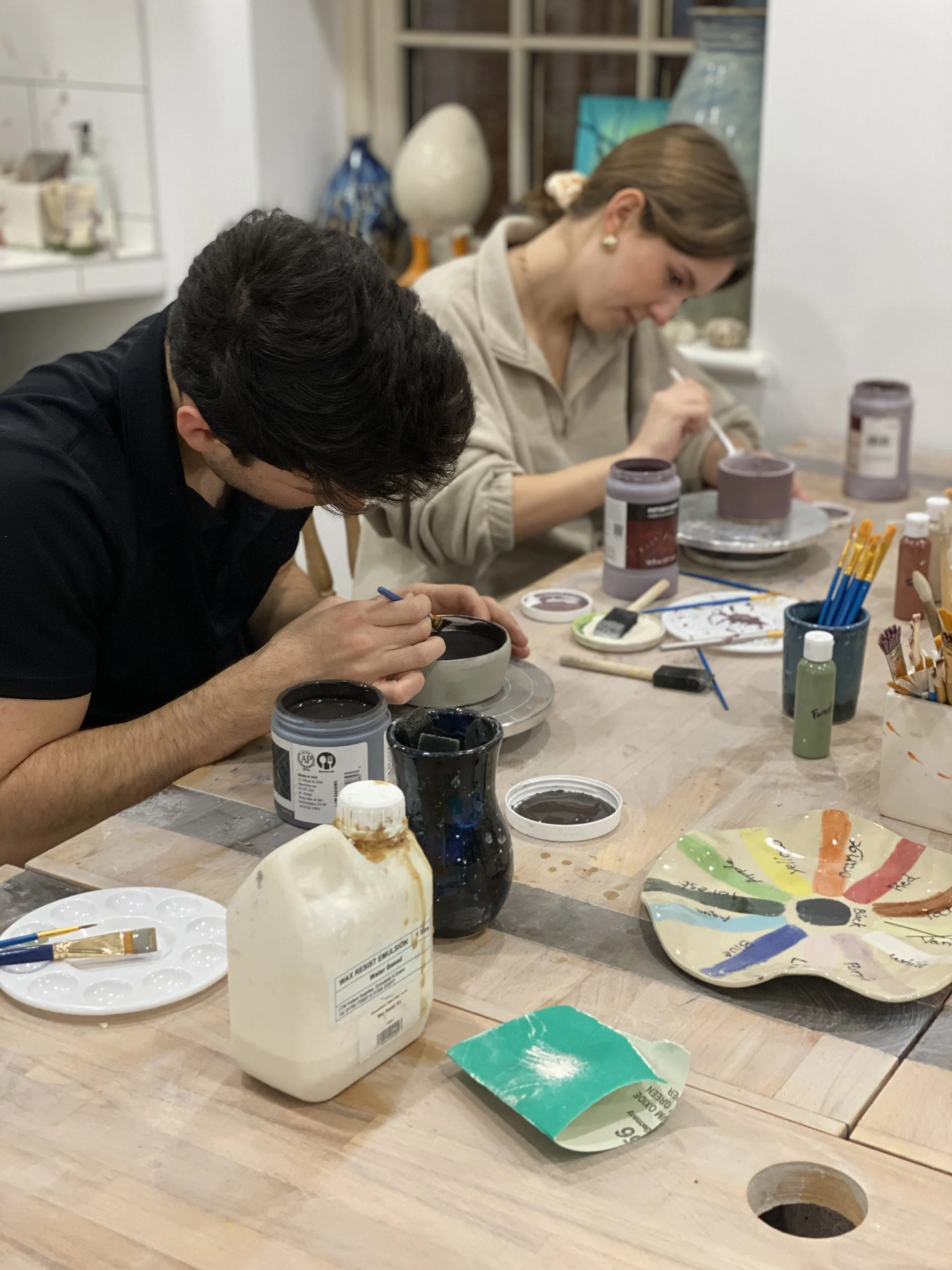 Two people painting pottery at a wooden table with various art supplies, including paint jars, brushes, and a color wheel.