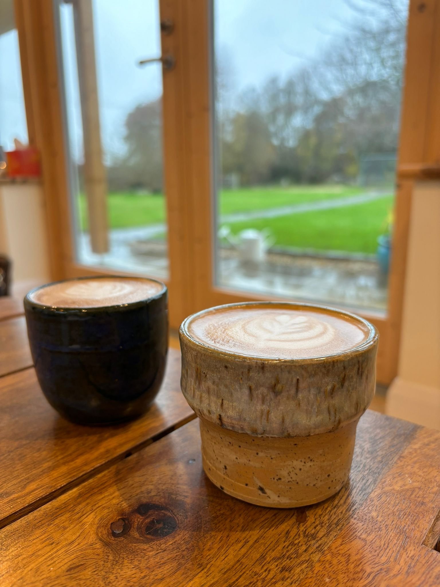 Two ceramic coffee cups with latte art on a wooden table near a large window showing a rainy outdoor scene with green grass and leafless trees.