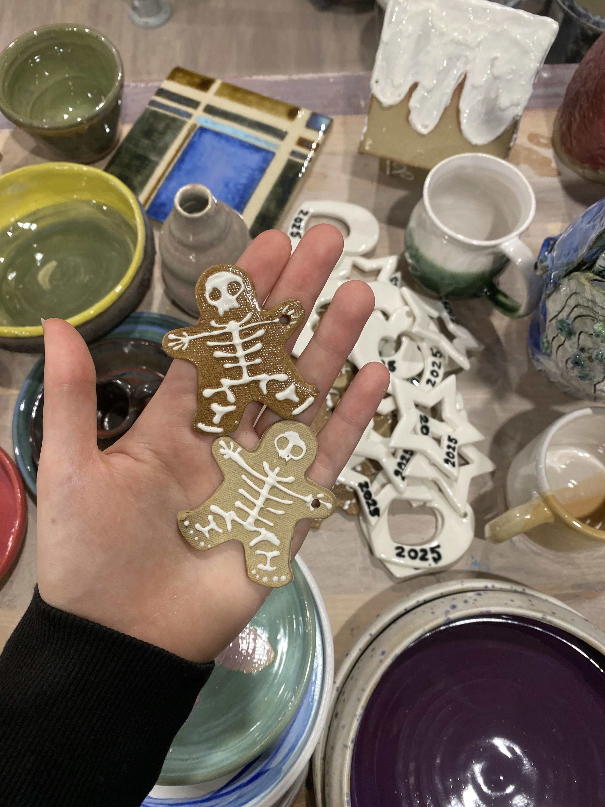 Person holding decorated gingerbread cookies shaped like skeletons with white icing detail, surrounded by various pottery and ceramic items on a tabletop.