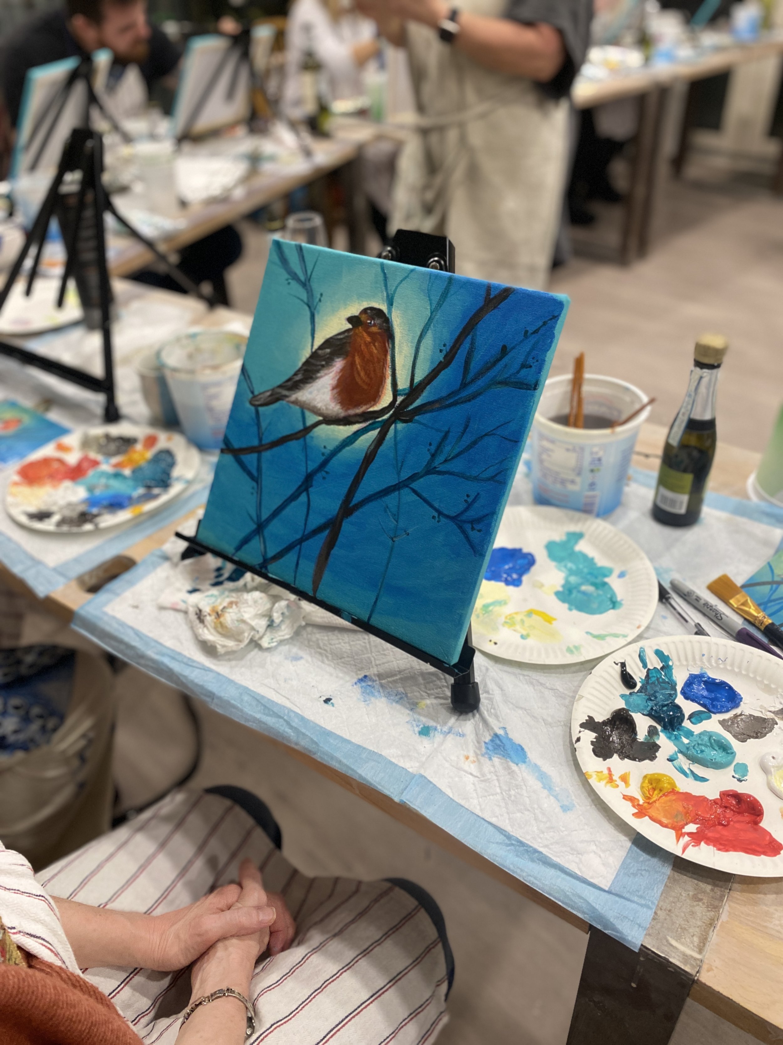 A painting of a bird on a branch, set up on a table during an art class. Art supplies and other paintings are visible in the background.