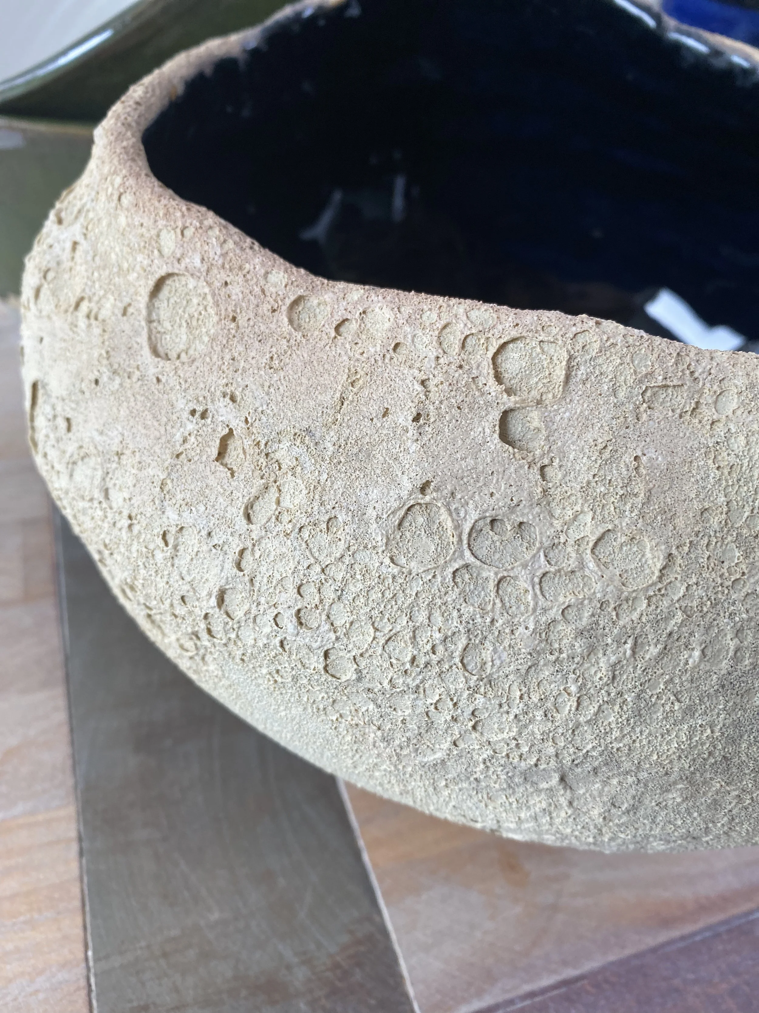 Close-up of a textured ceramic bowl with a rough, bubbly surface exterior and a dark interior, on a wooden surface.