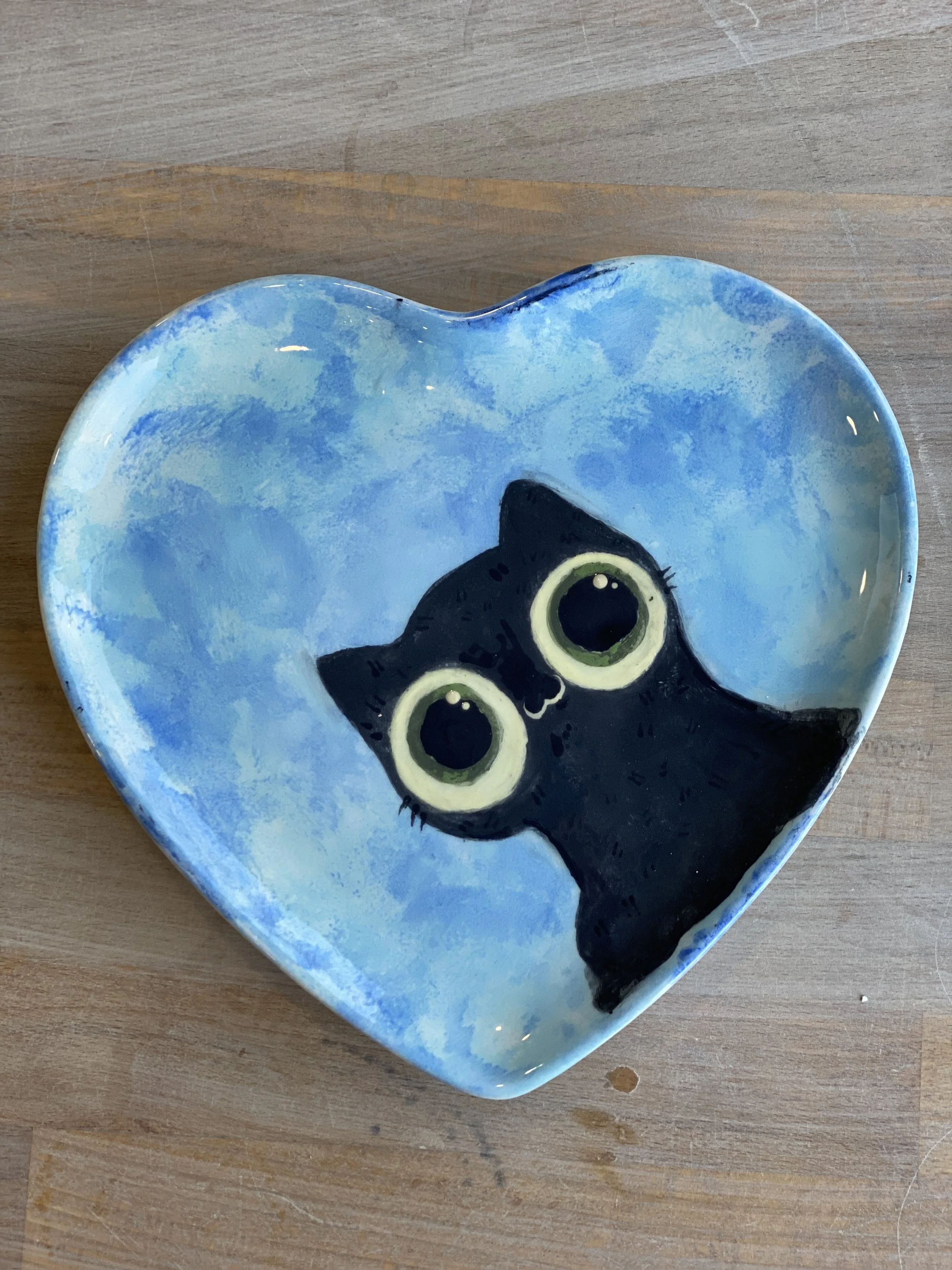 A heart-shaped ceramic dish with a painting of a black cat with large, green eyes, set against a blue background.