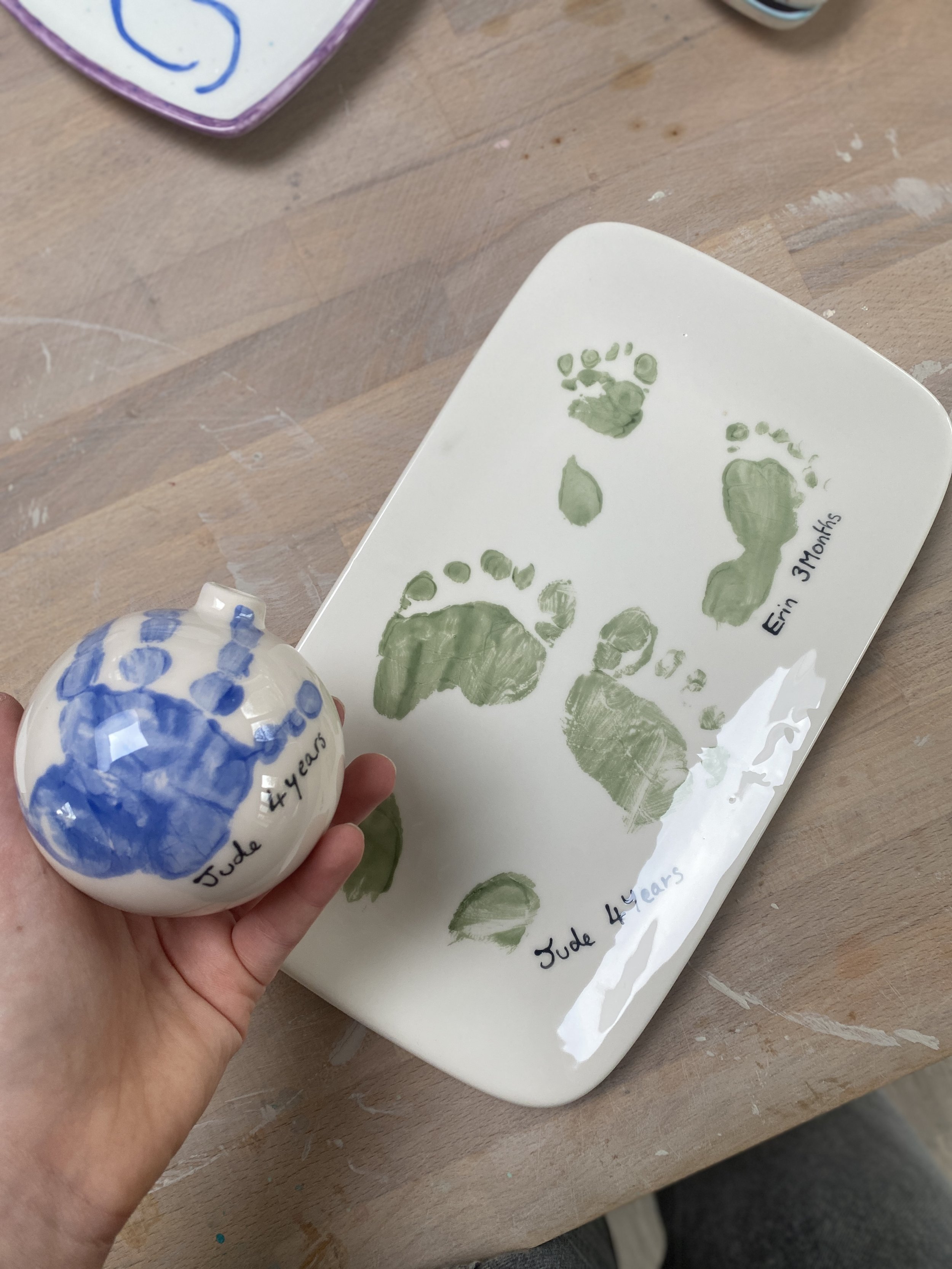A small white ornament with blue paint, labeled 'Jude 4 years', held in a person's hand. A white tray with green painted footprints and handprints, labeled 'Jude 4 years,' 'Erin 3 months' on a wooden surface.