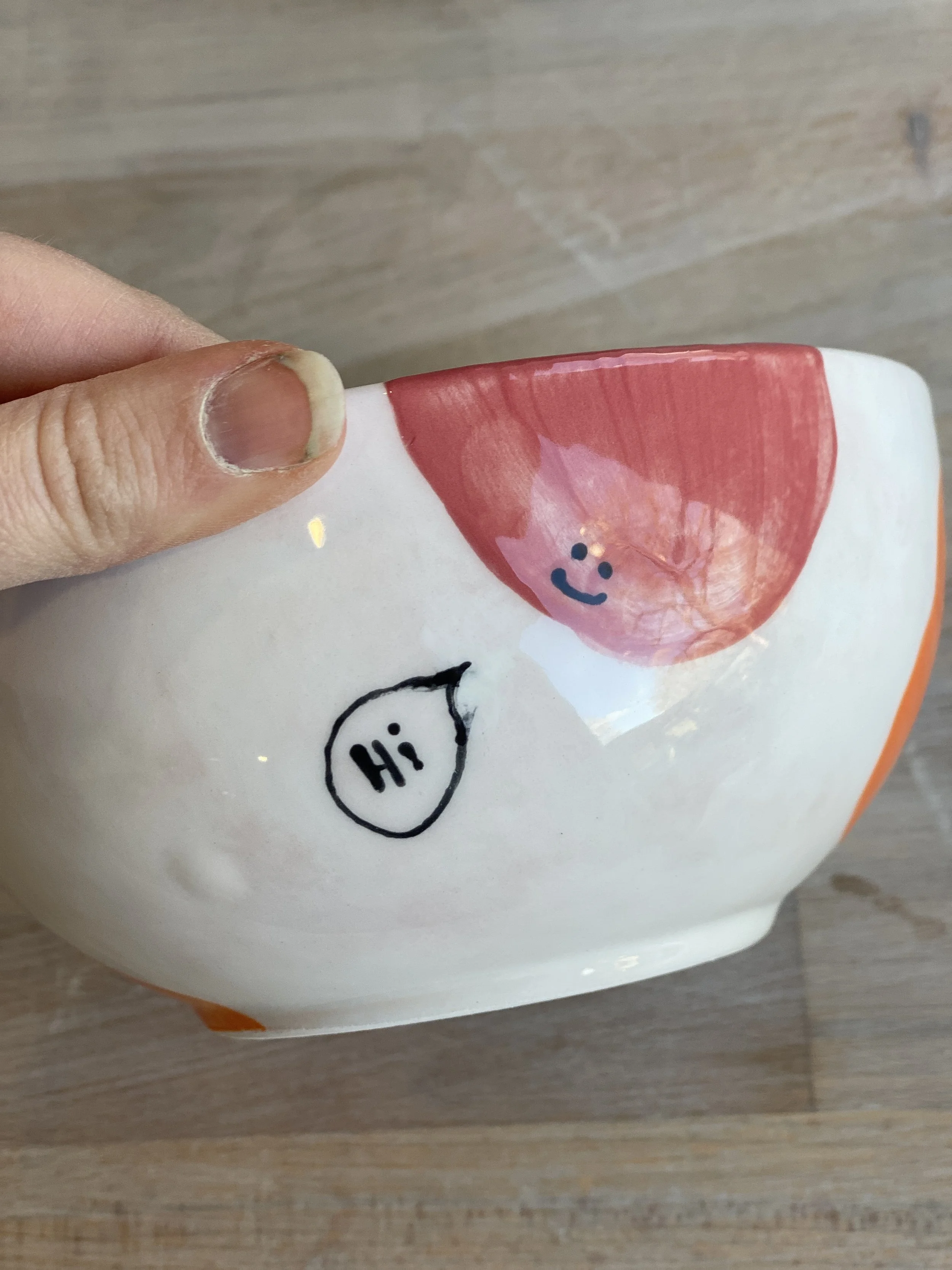 Close-up of a ceramic cup with hand-drawn illustrations, one saying 'Hi' inside a speech bubble and a smiling face with pink hair.