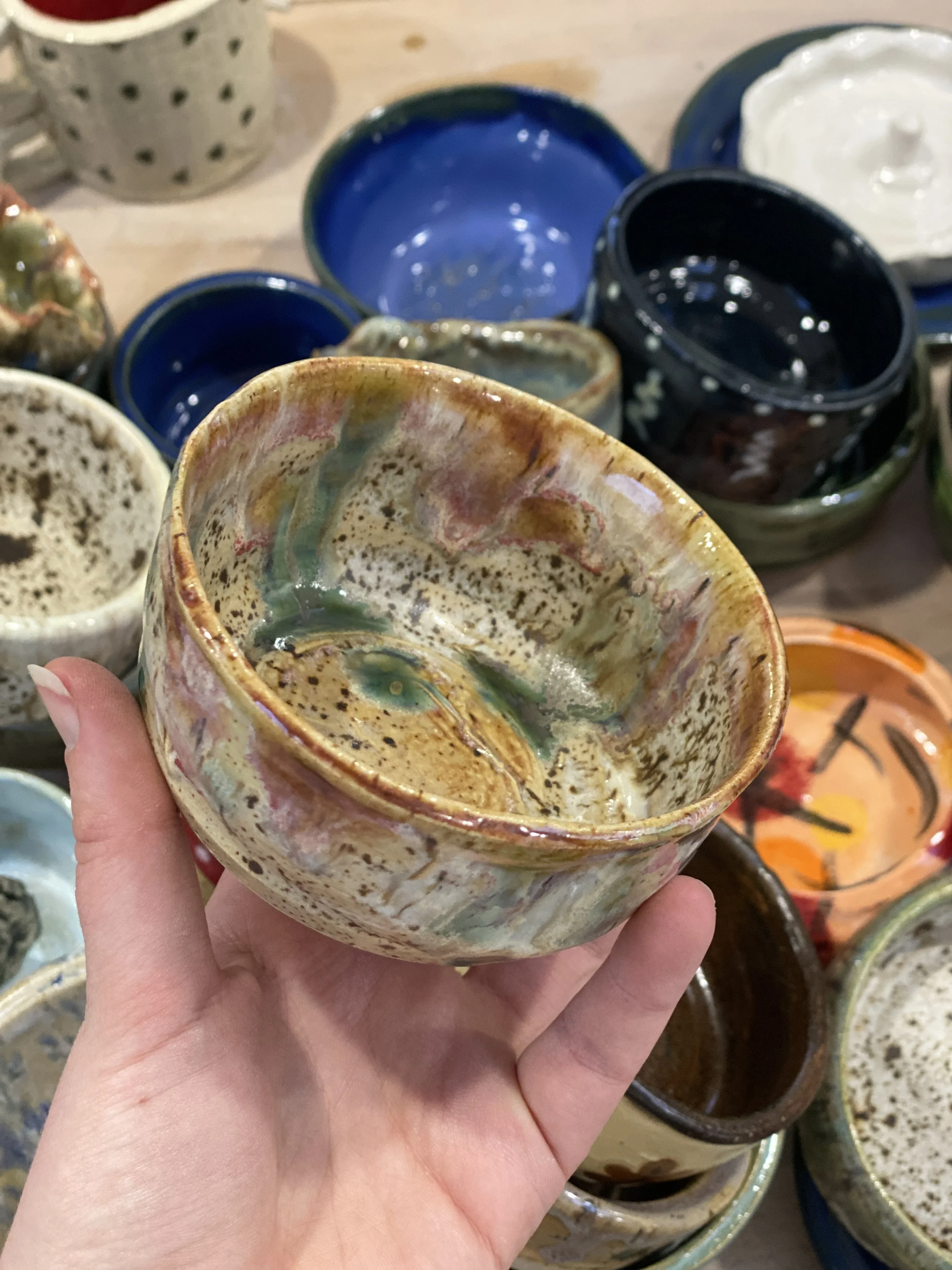 Hand holding a speckled, multicolored ceramic bowl among various other bowls with colorful glazes on a table.