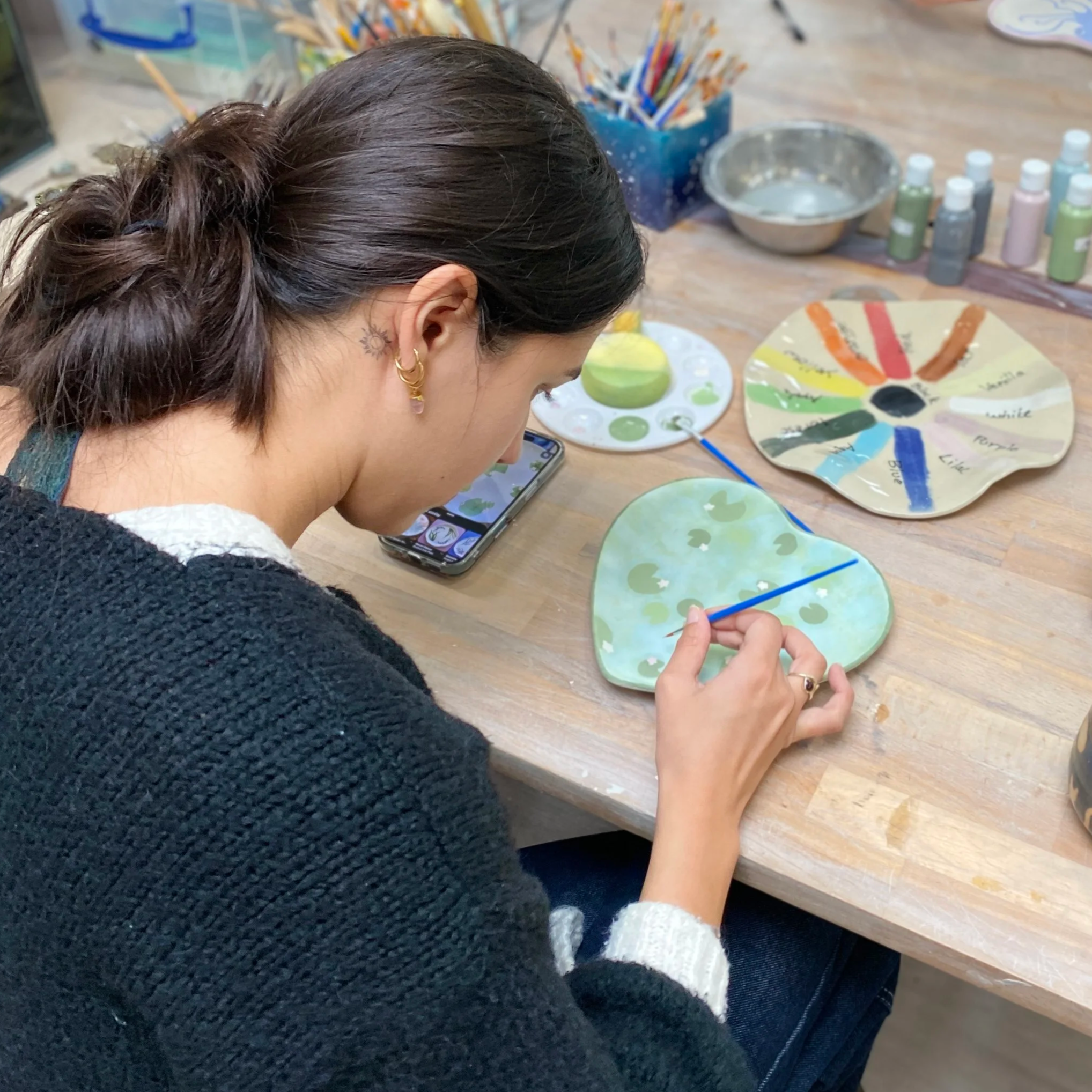 A woman with dark hair, gold earrings, and a tattoo of a sun behind her ear, painting ceramic plates with a small brush at a wooden table. There are colorful paint bottles, a color wheel, and other art supplies on the table.