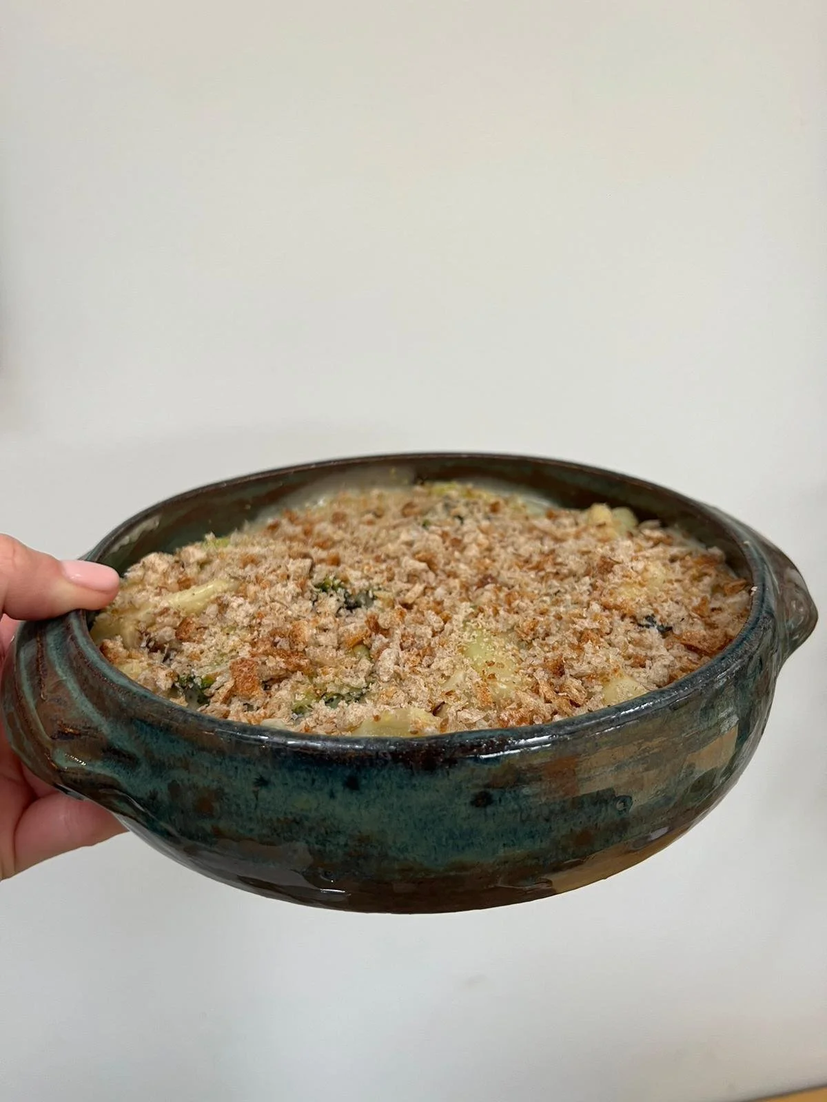 A person holding a ceramic dish filled with baked casserole topped with breadcrumbs, cheese, and herbs against a plain white background.