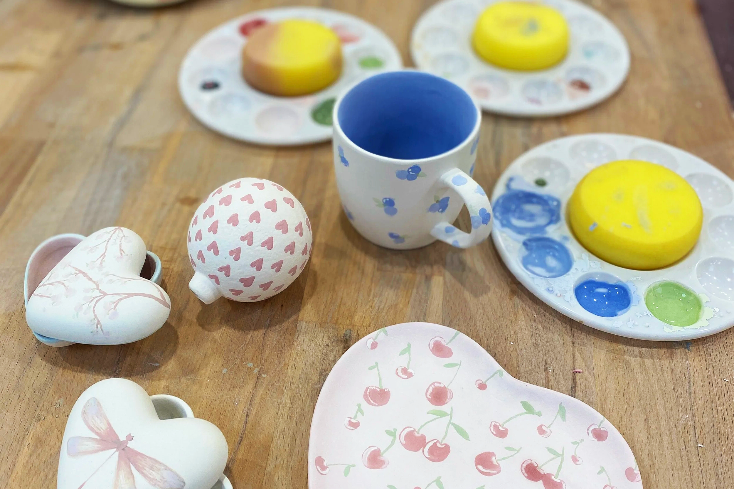 Painted ceramic objects including two heart-shaped pieces, a spherical object with pink hearts, a cup with blue flowers, and several painted plates with food-themed designs on a wooden table.