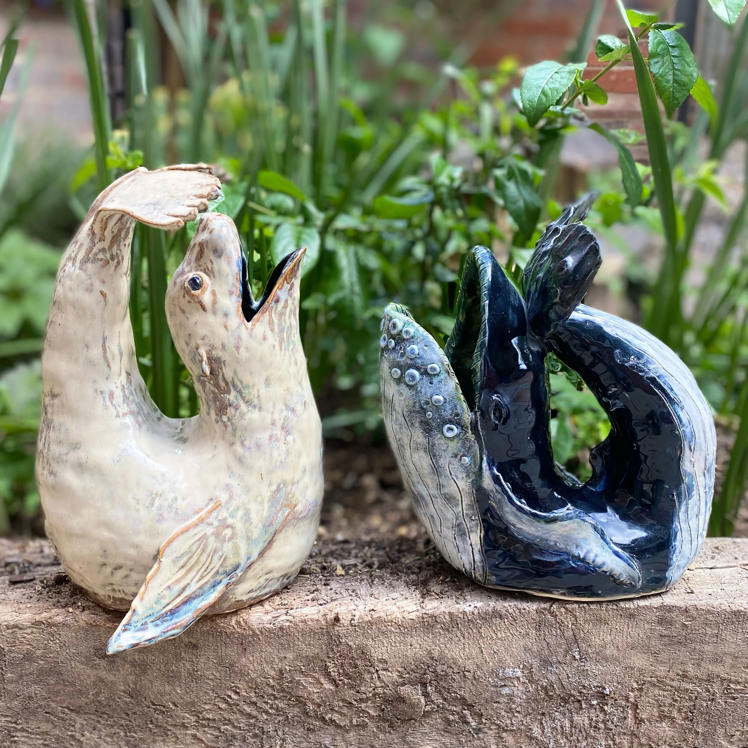 Two ceramic bird sculptures, one light-colored and the other dark-colored, are placed on a wooden surface outdoors among green plants.