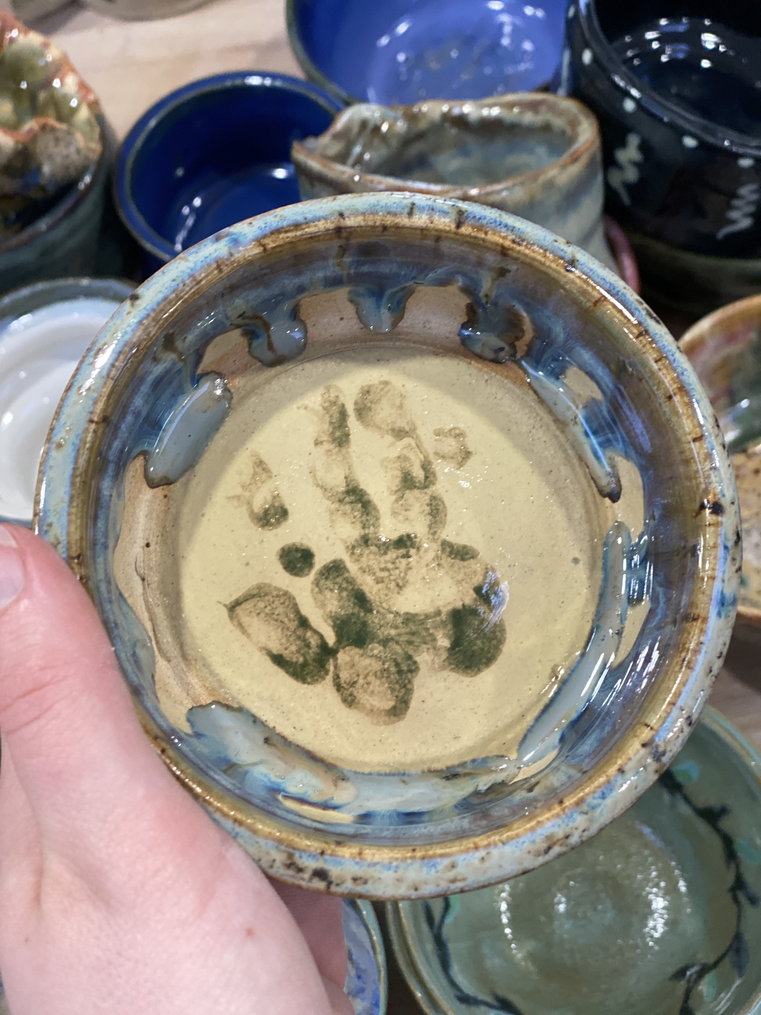 A person holding upside-down ceramic bowl with a glaze finished surface, blue and brown accents, surrounded by other glazed ceramic bowls and cups.