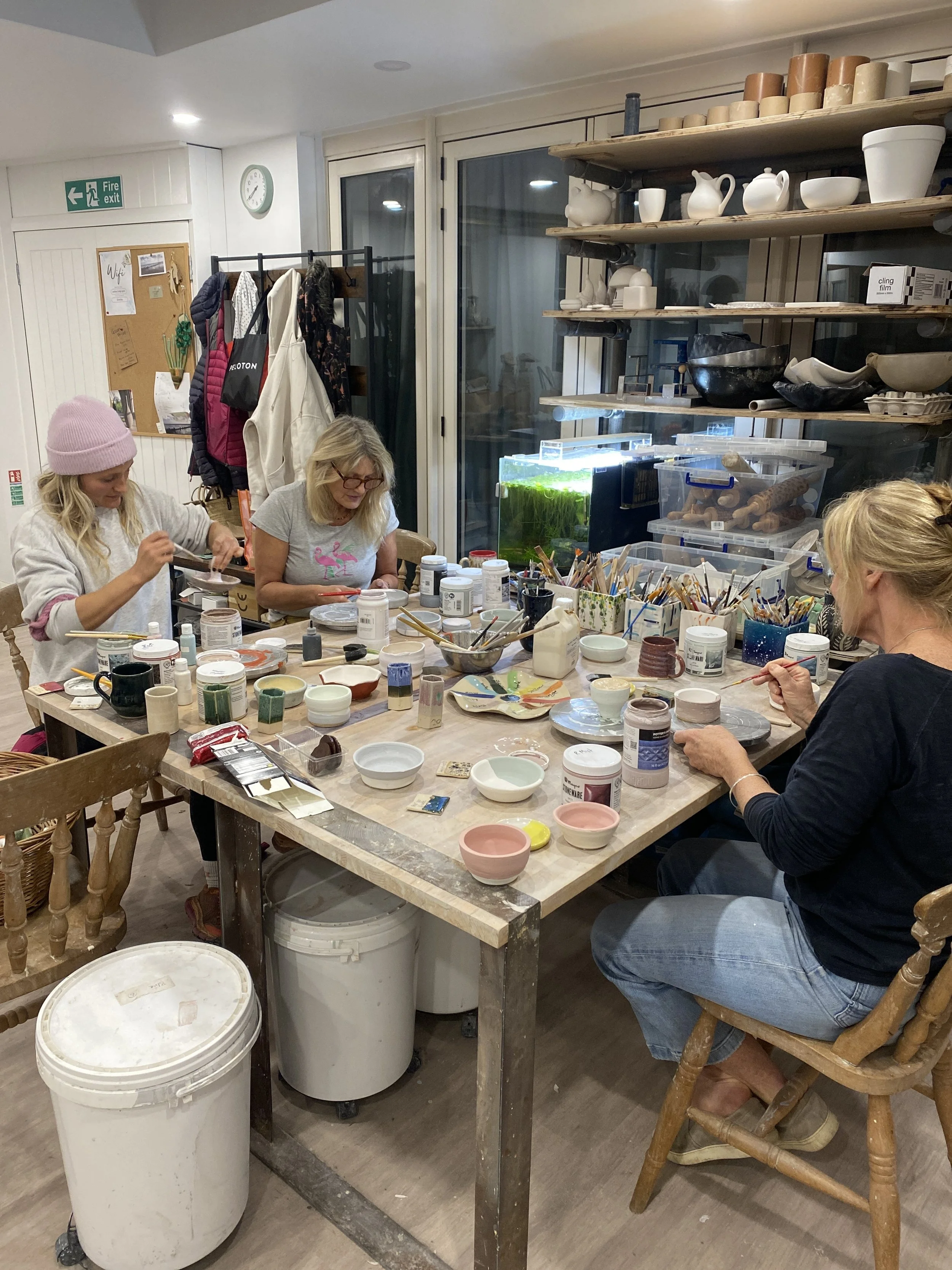 Three women are painting ceramic pottery at a craft workshop table with various paint supplies and ceramics. The setting appears to be a ceramics studio with shelves of pottery and art supplies in the background.